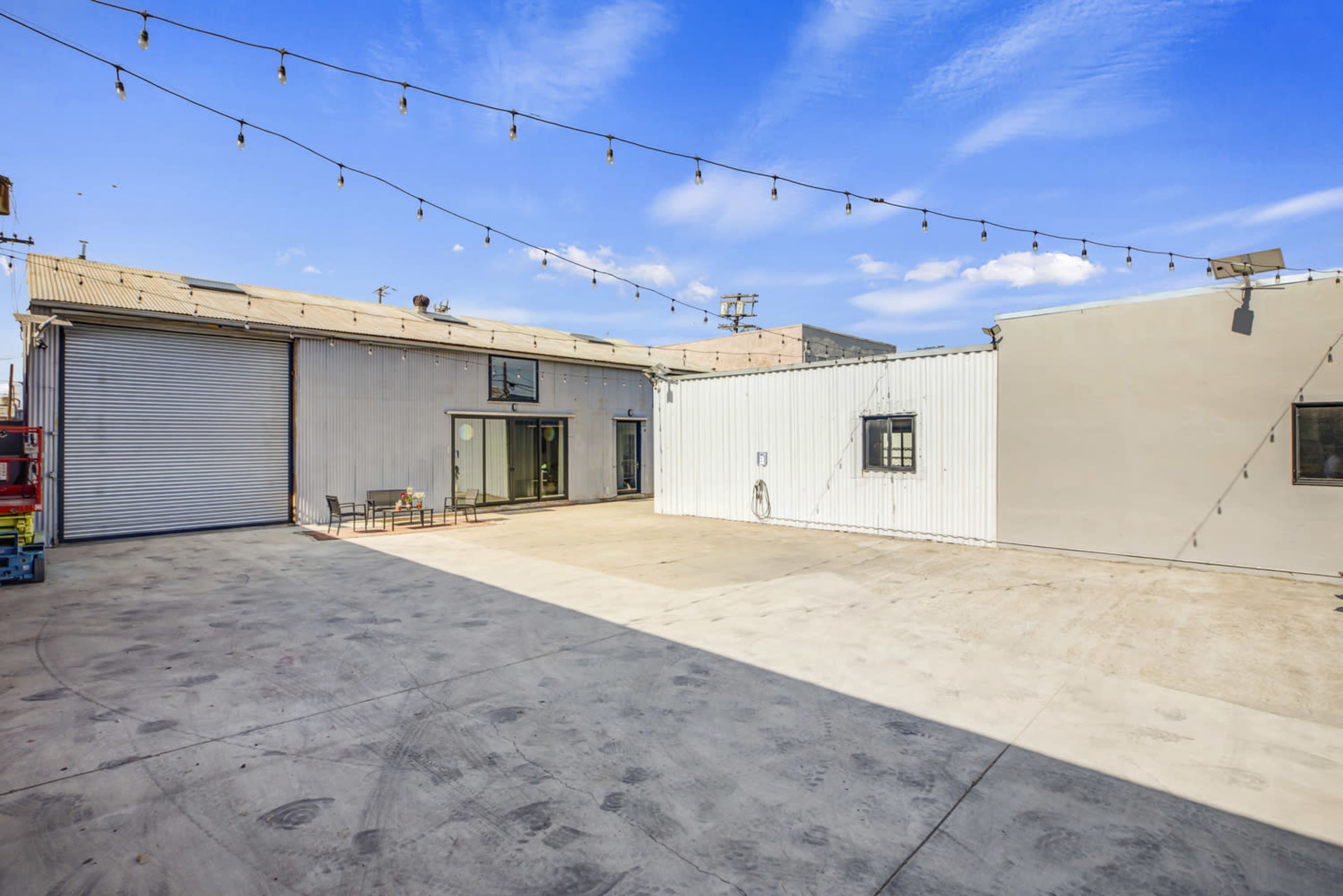The image shows a spacious outdoor area with a concrete floor, flanked by industrial buildings and string lights overhead.