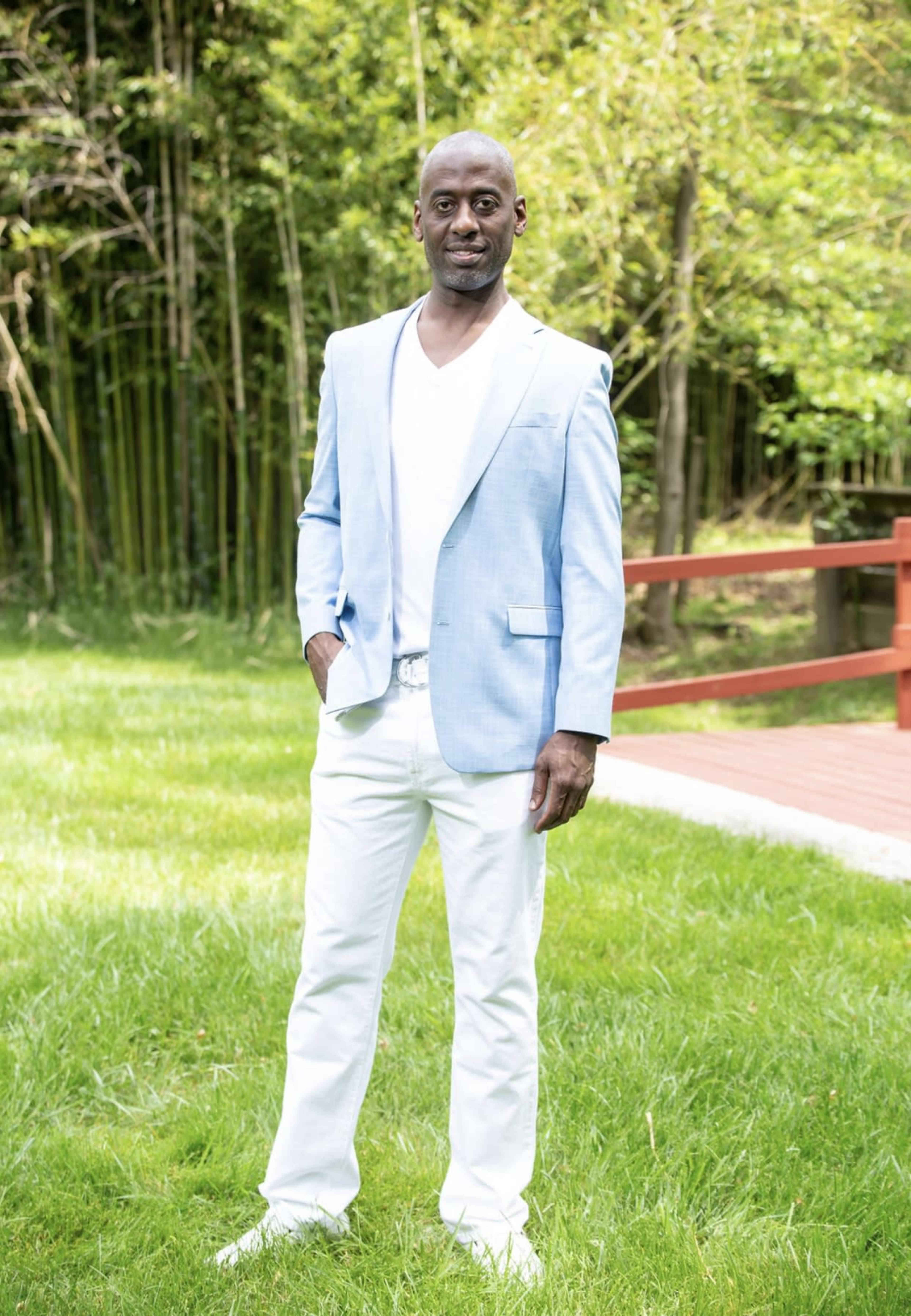 A man stands on a grassy area near a wooden deck, dressed in a light blue blazer, white shirt, and white pants.