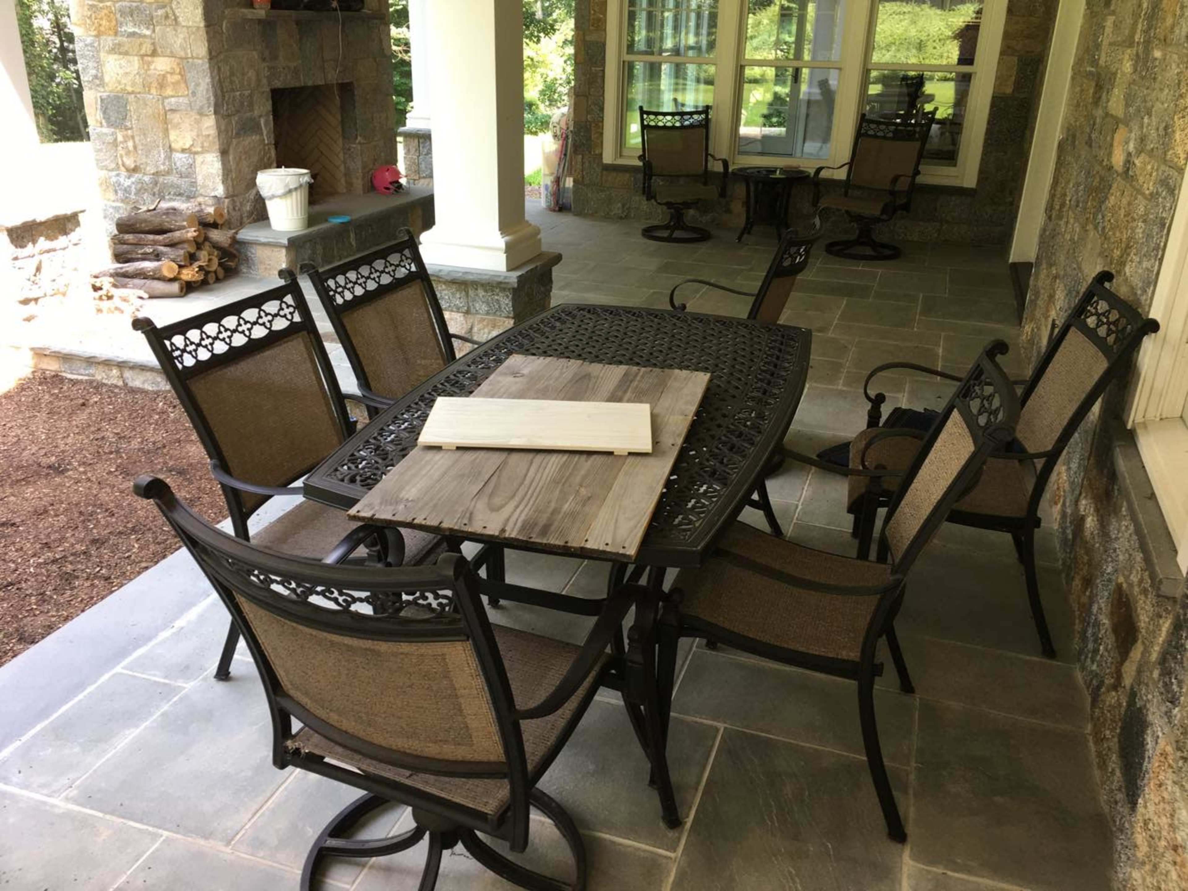 A patio area features a metal dining table surrounded by several chairs, with a wooden board placed on top of the table.