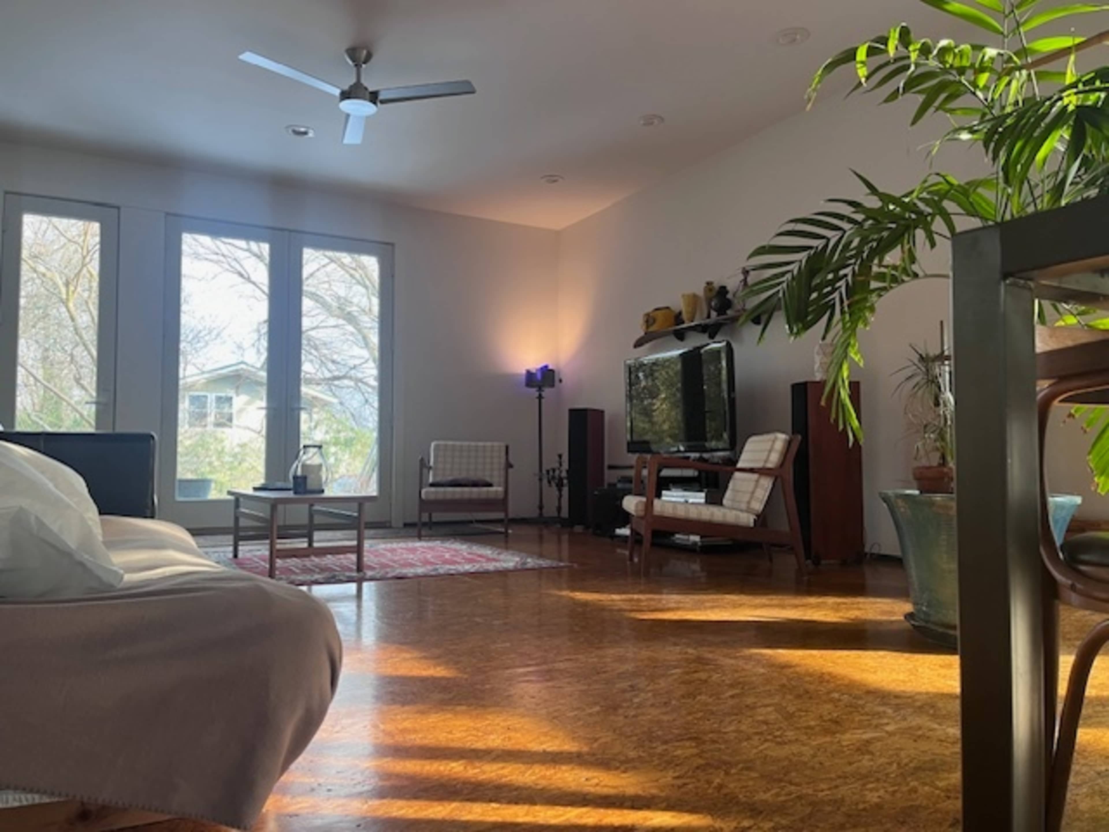 A well-lit living room features a couch, two chairs, a television stand, and several indoor plants.