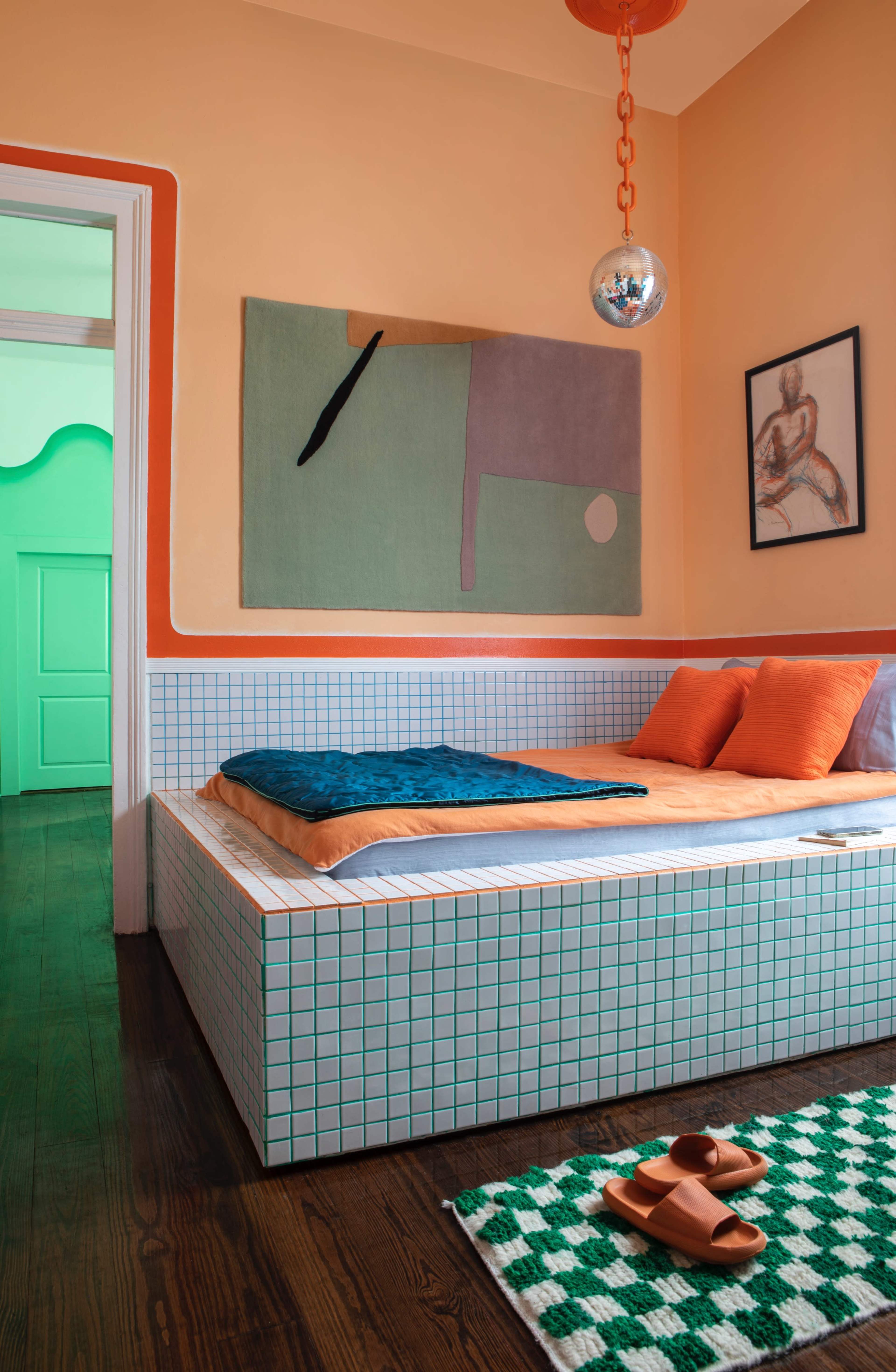 The image features a modern bedroom with a tiled bed frame, orange walls, and colorful decor, including a large artwork and a round mirror pendant.