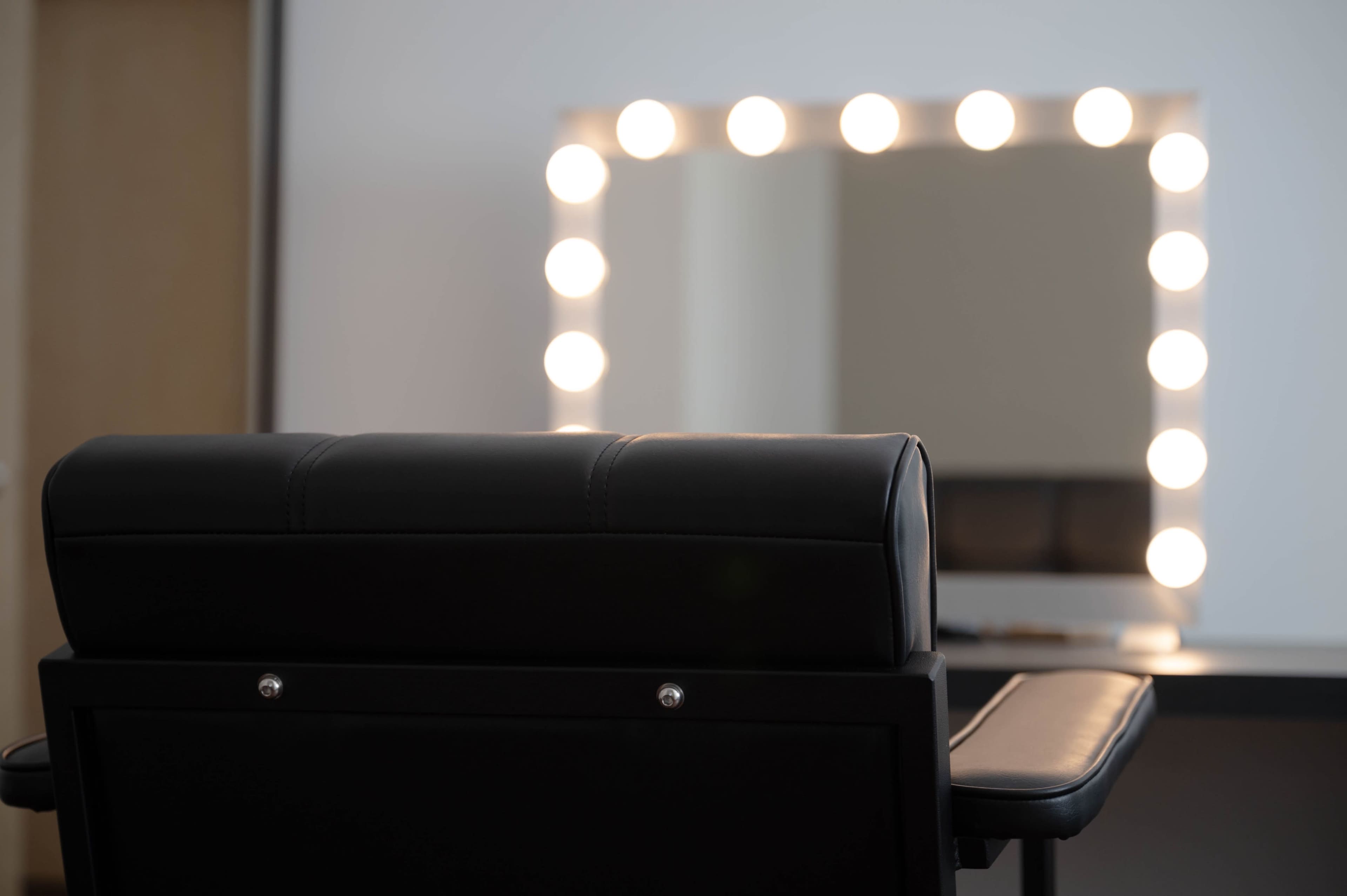 A black salon chair is positioned in front of a well-lit mirror with circular lights.