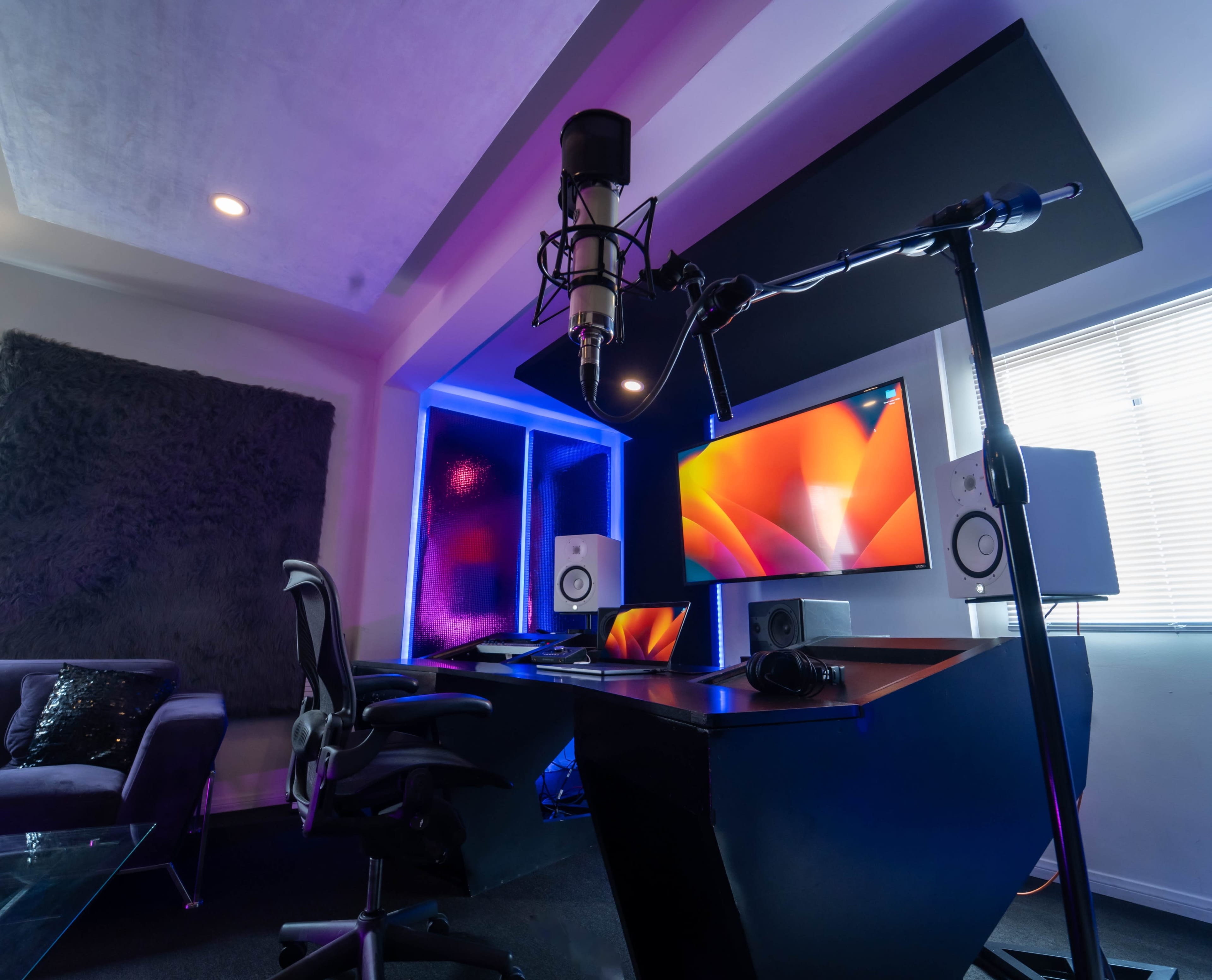 A recording studio setup featuring a microphone on a stand, a computer monitor, speakers, and an office chair arranged around a modern desk.