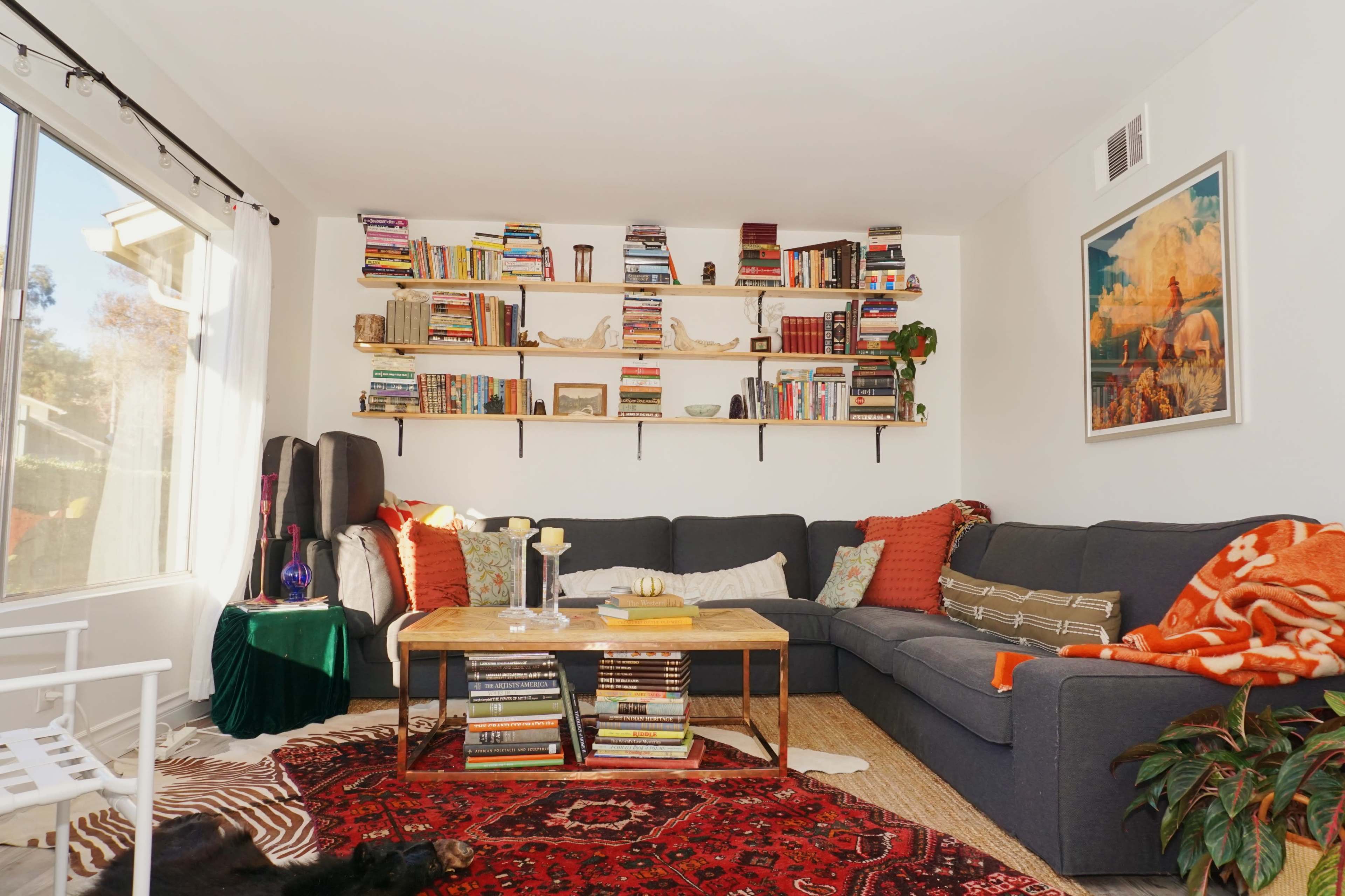 A cozy living room features a large sectional sofa adorned with colorful cushions, surrounded by bookshelves filled with books and decorative items.