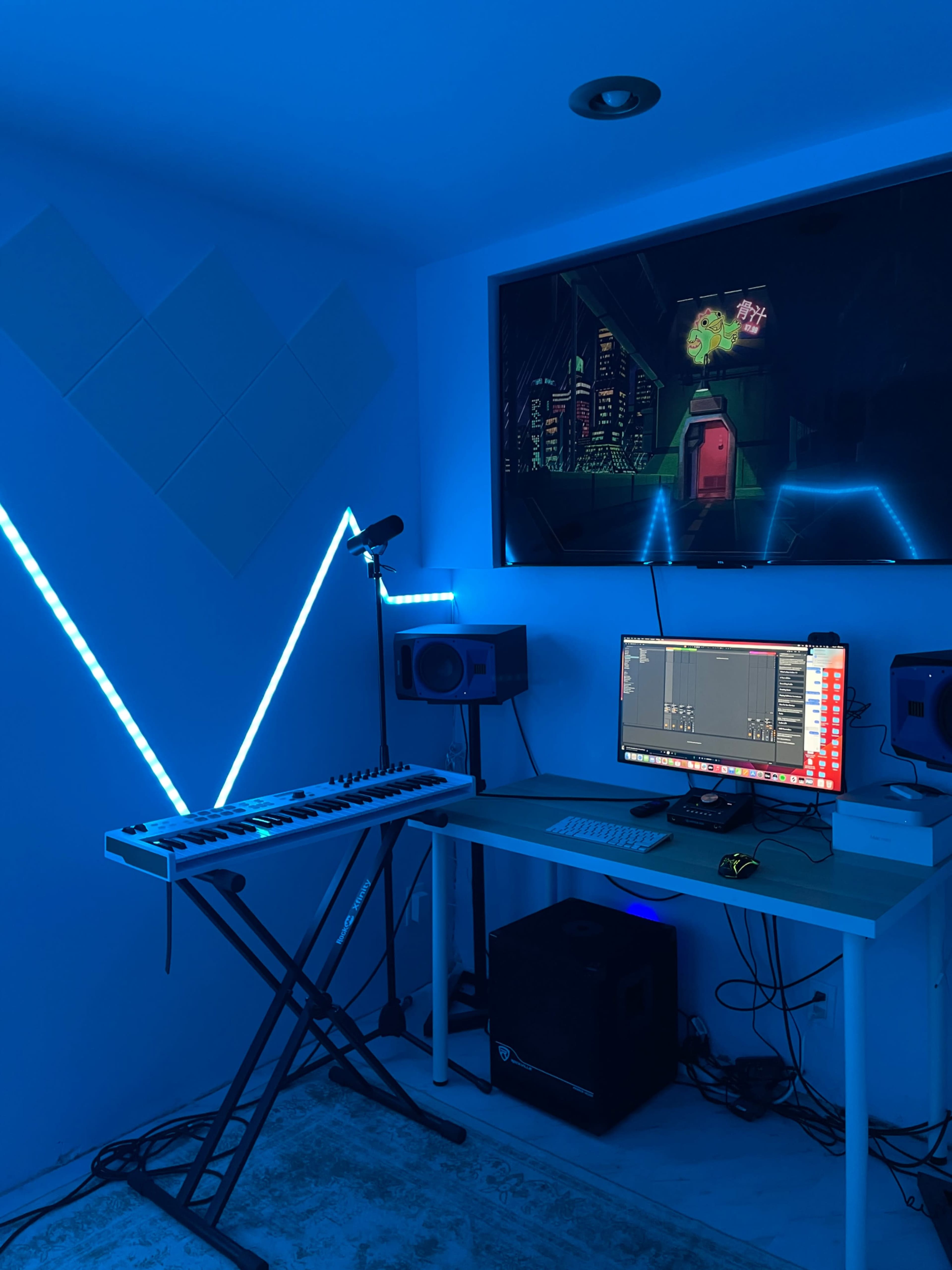 A music production setup features a keyboard, computer, and speakers under blue ambient lighting, with a digital artwork displayed on the wall-mounted screen.
