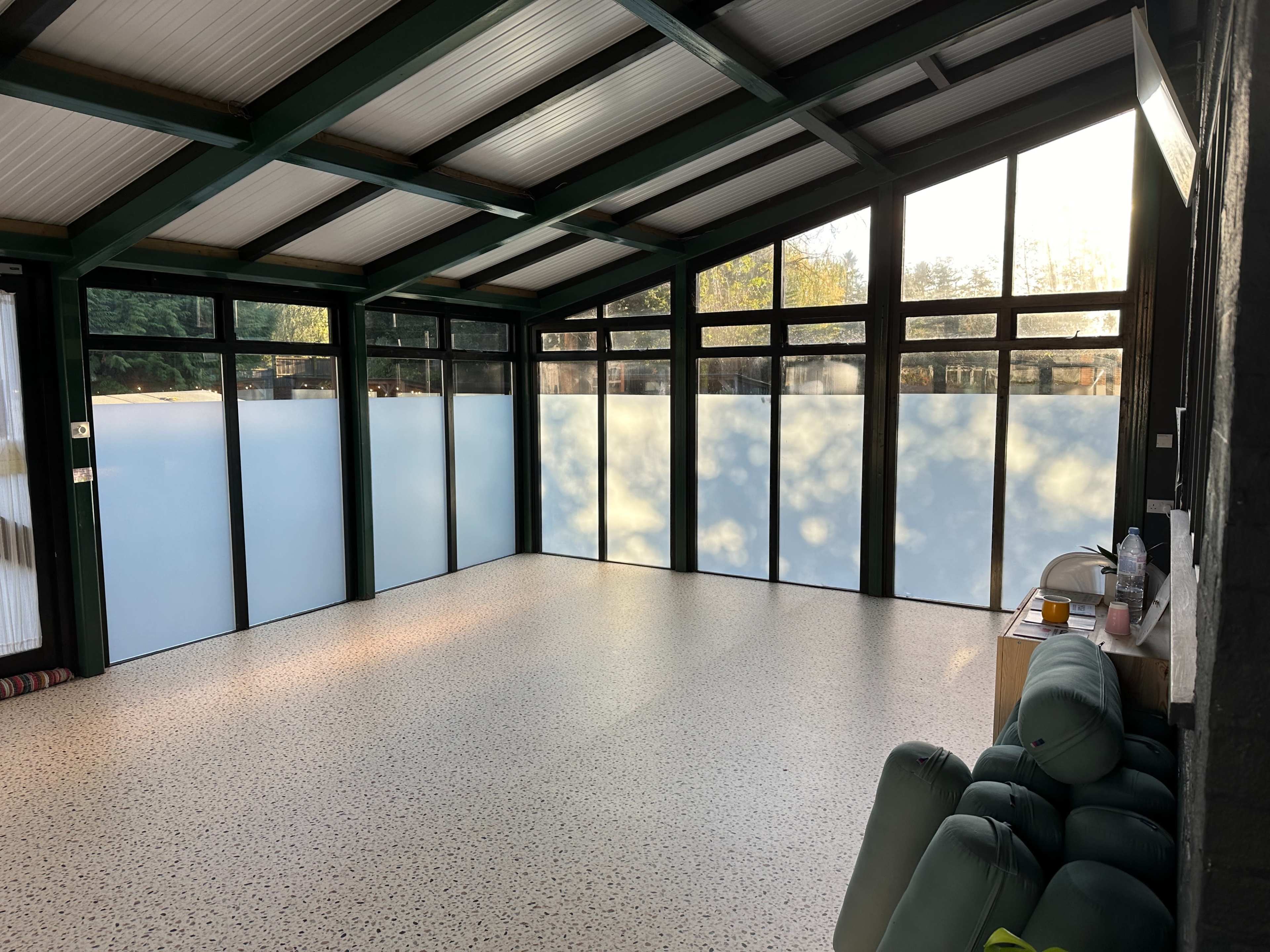 The image shows a spacious, modern room with large windows and a smooth floor, featuring green beams and minimal furnishings.