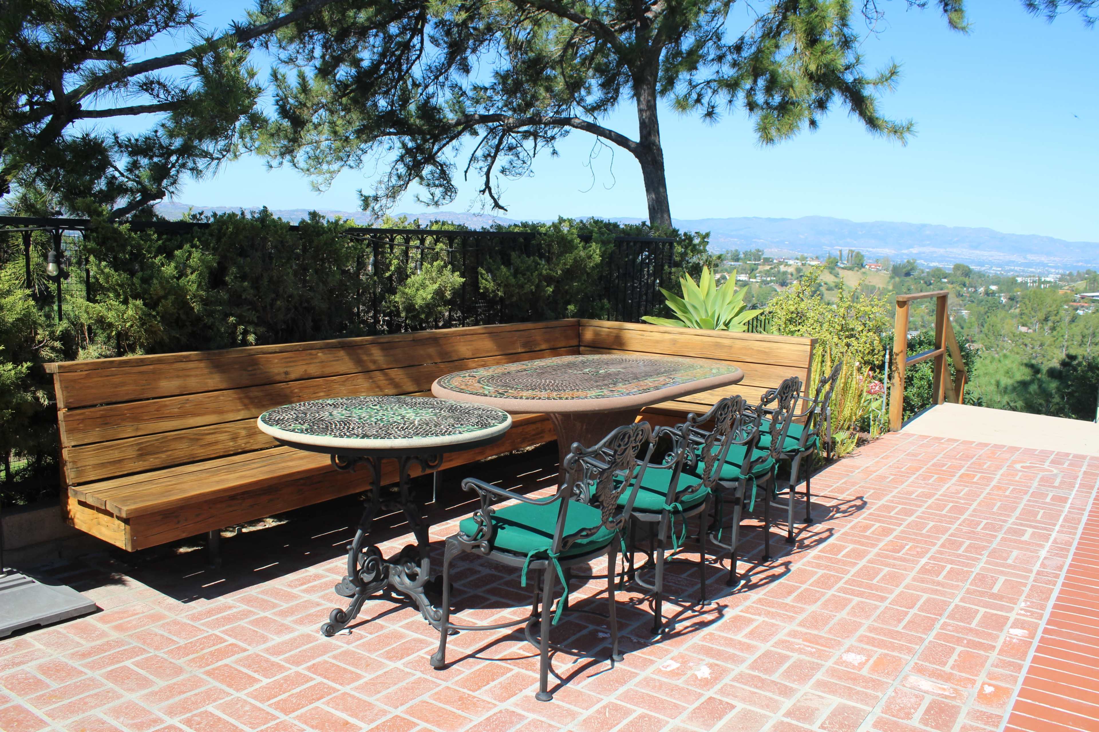 A wooden bench and a set of metal and green cushioned chairs surround a round table on a brick patio.