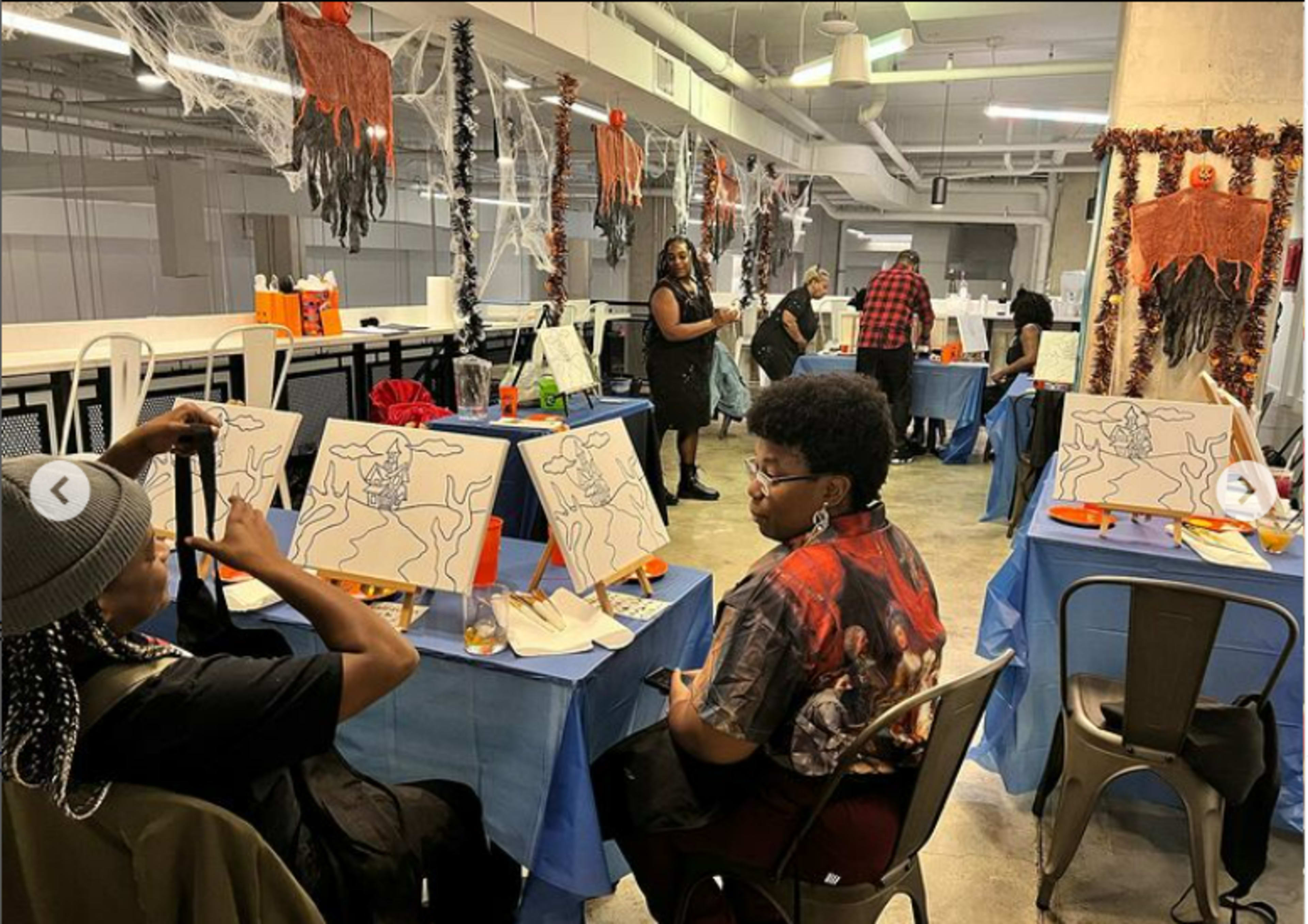 Two individuals sit at a table adorned with canvases and art supplies in a decorated room featuring Halloween-themed decorations and people engaged in painting activities.