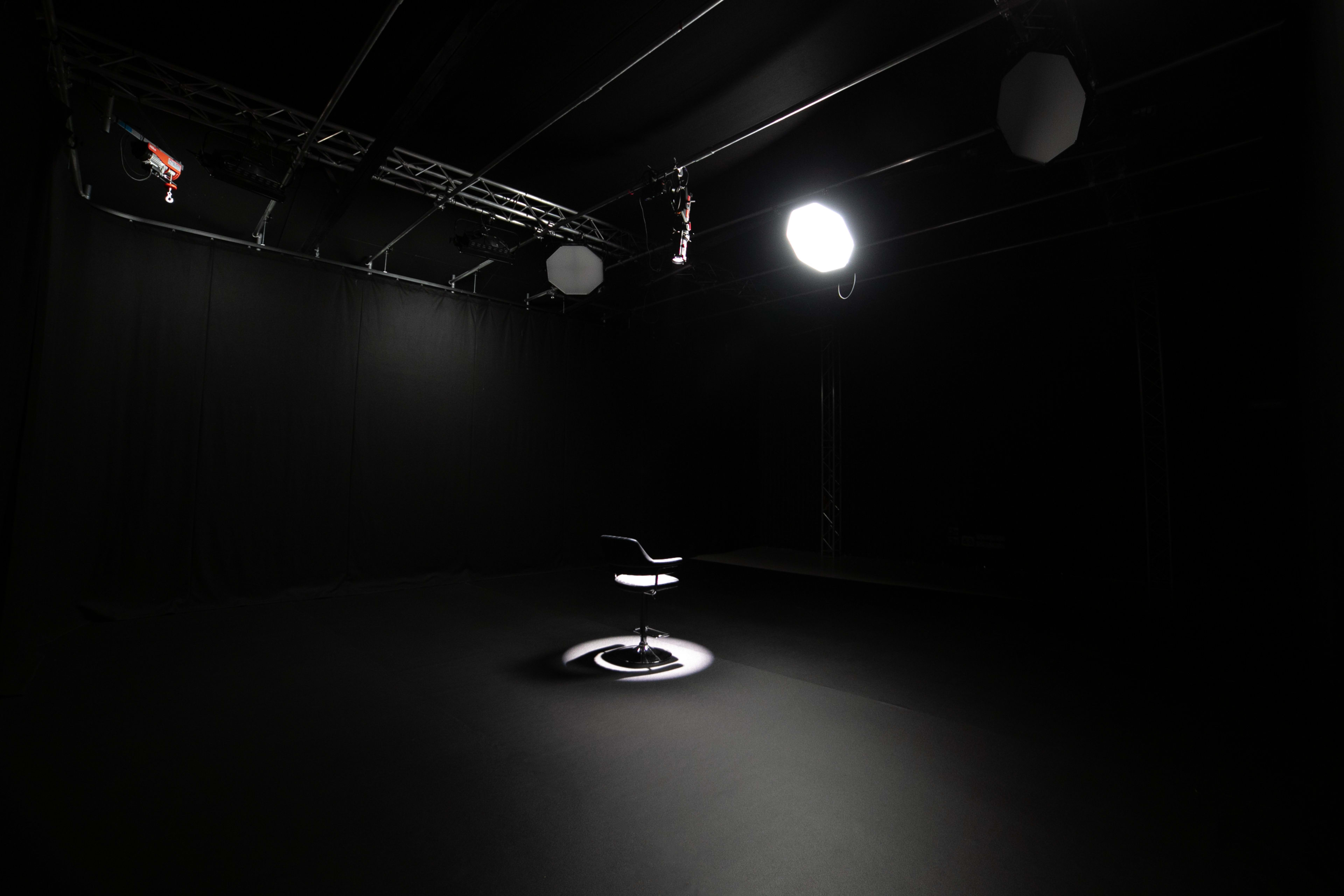 The image shows a dimly lit studio space with a single chair in the center, illuminated by a spotlight against a black backdrop.