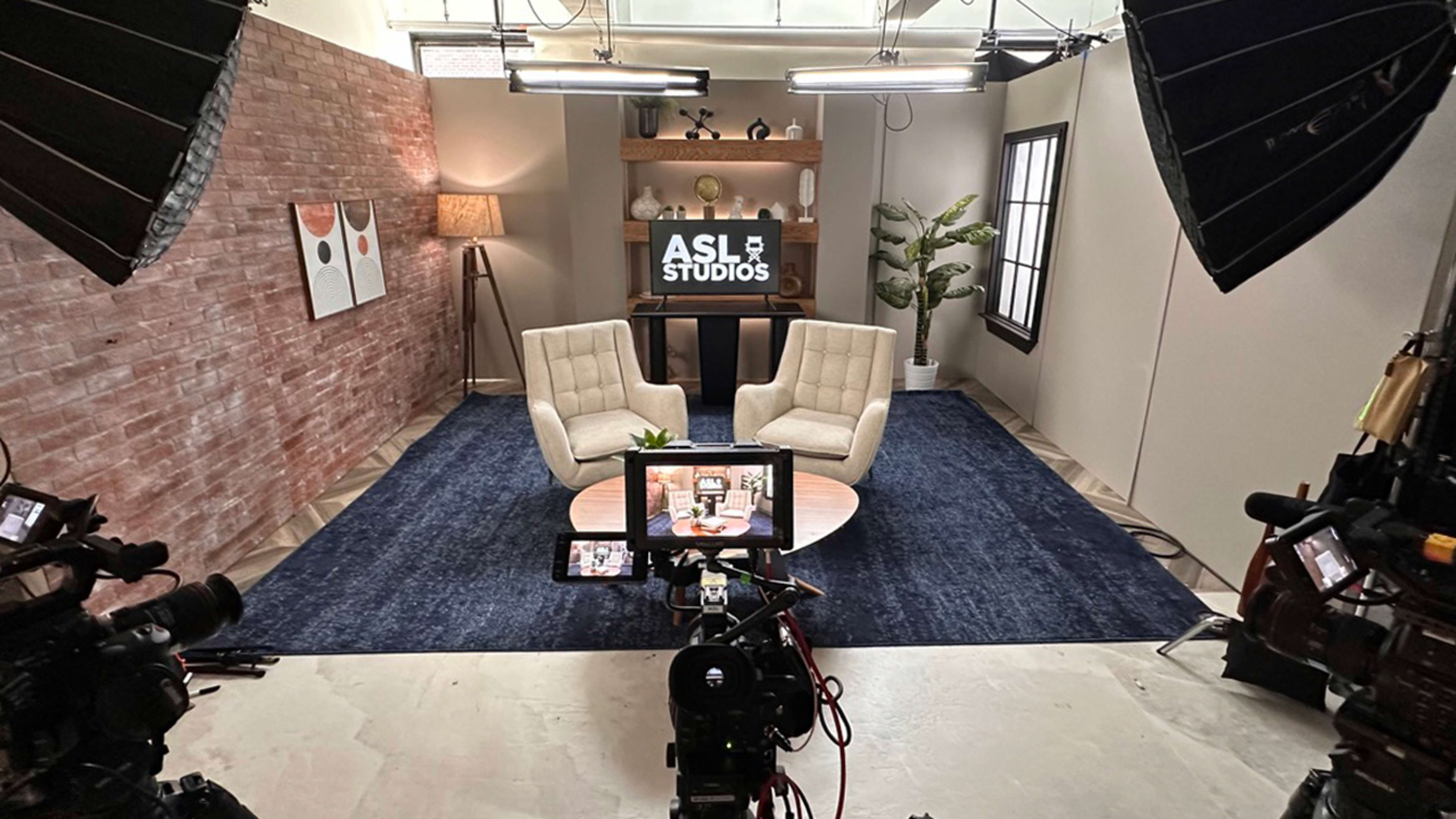 The image shows a television studio setup with two chairs, a coffee table, and a backdrop featuring the text "ASL Studios" on a screen, surrounded by lights and camera equipment.