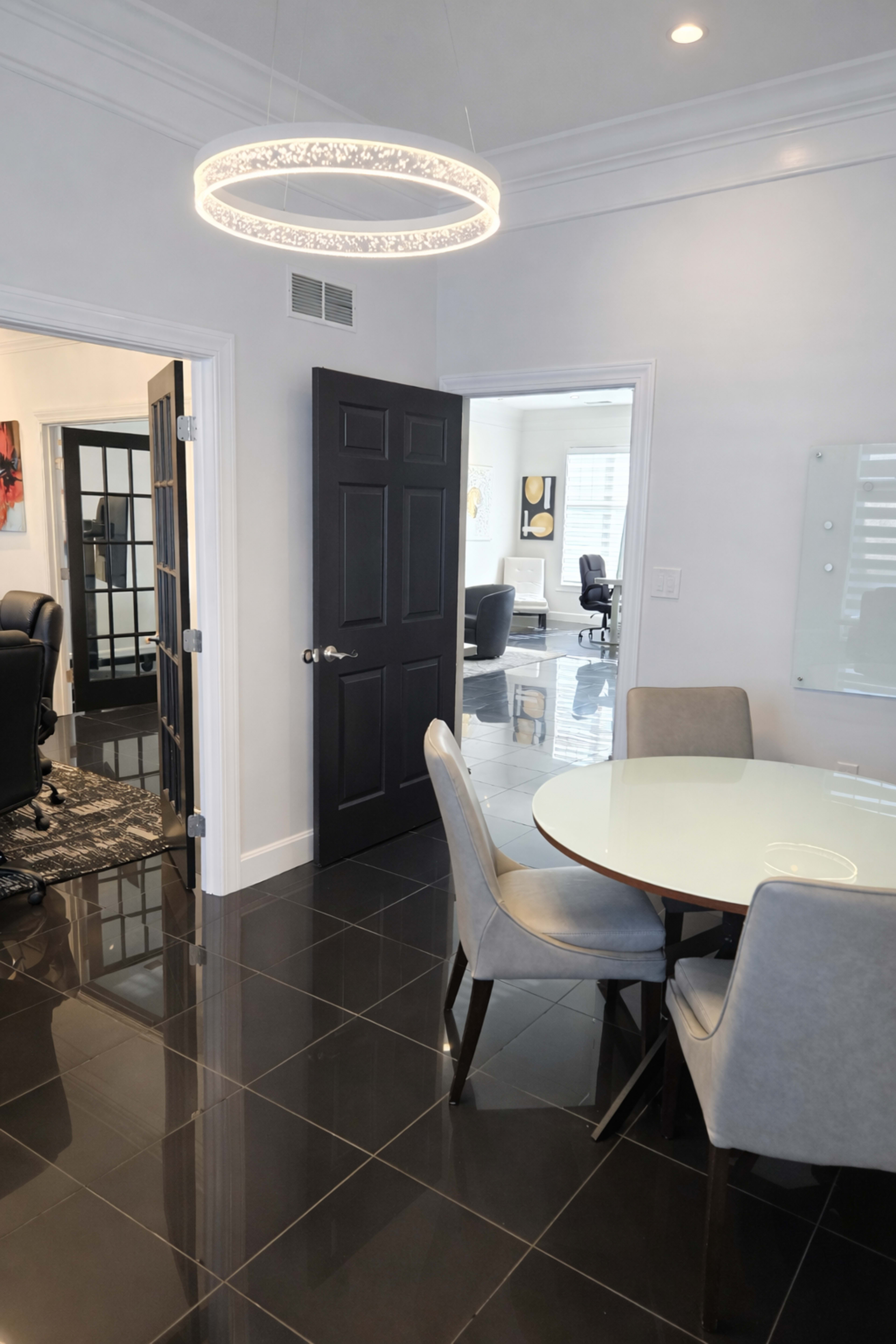The image shows a modern office space featuring a round dining table with gray chairs, black tiled flooring, and a circular light fixture, with a doorway leading to another room in the background.