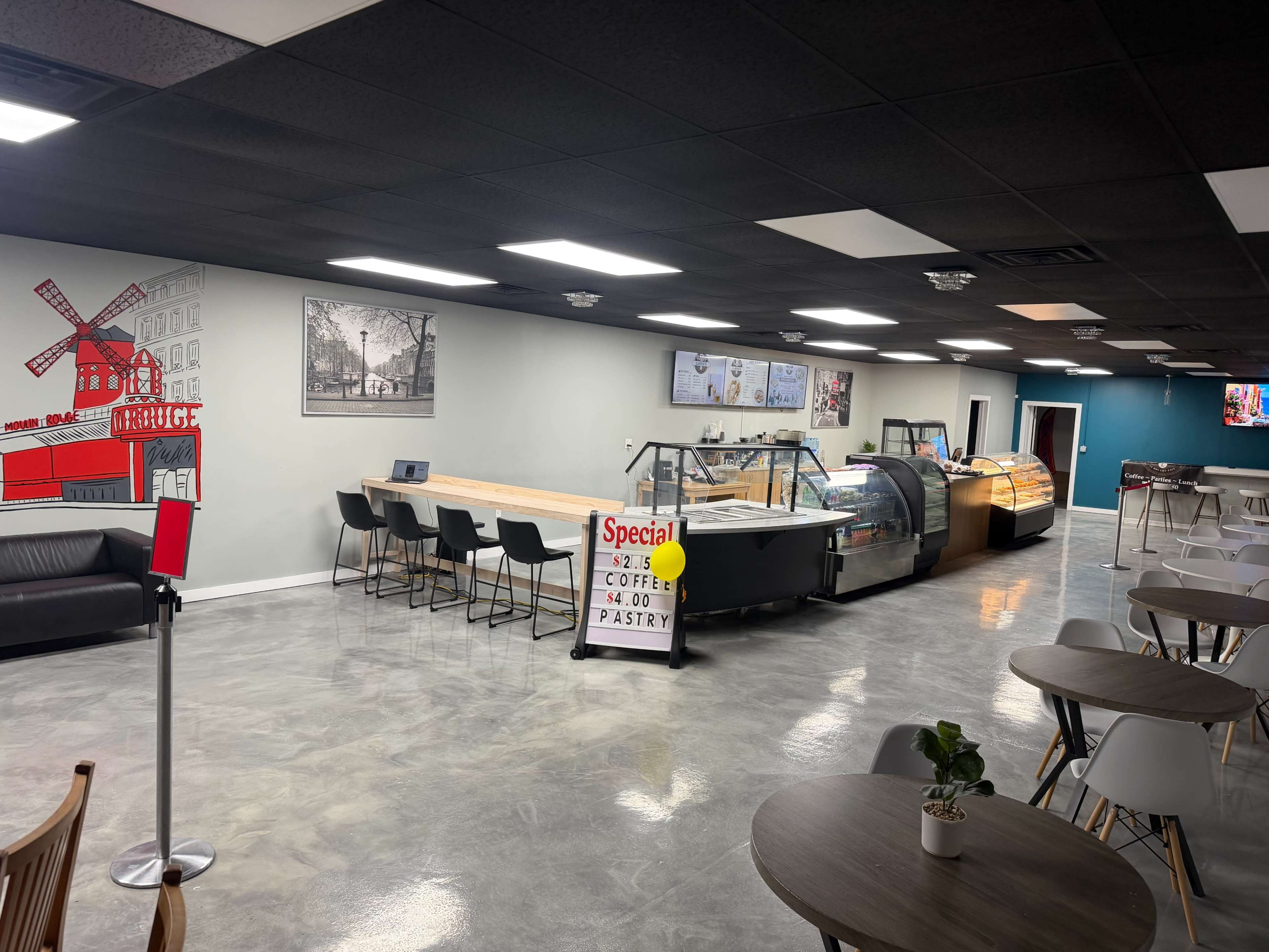 The image shows a modern café interior with a display case, seating areas, and promotional signage for coffee and pastry specials.