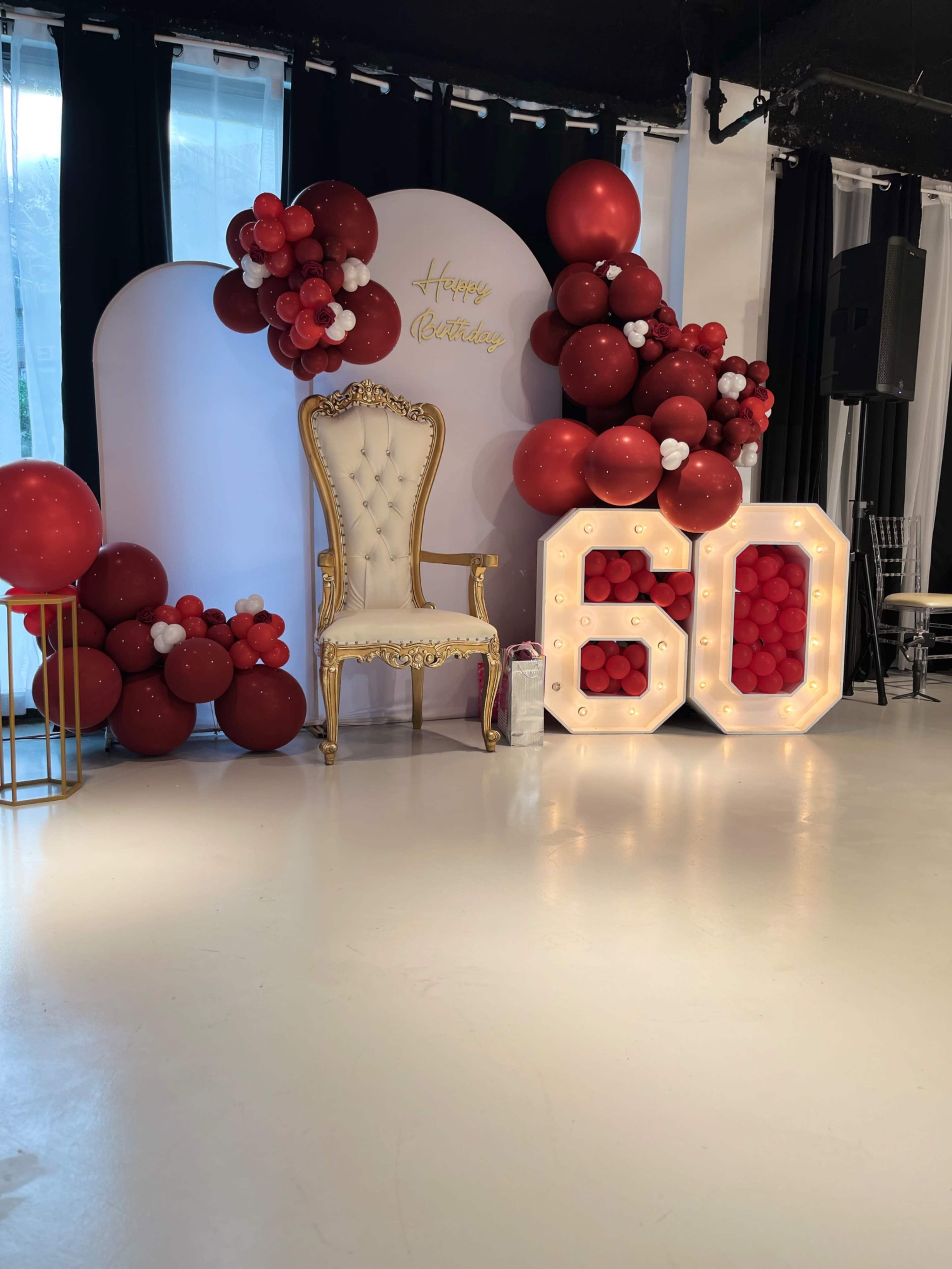 A decorated stage featuring a golden chair, a "Happy Birthday" sign, and large illuminated numbers "60" alongside red and white balloon arrangements.