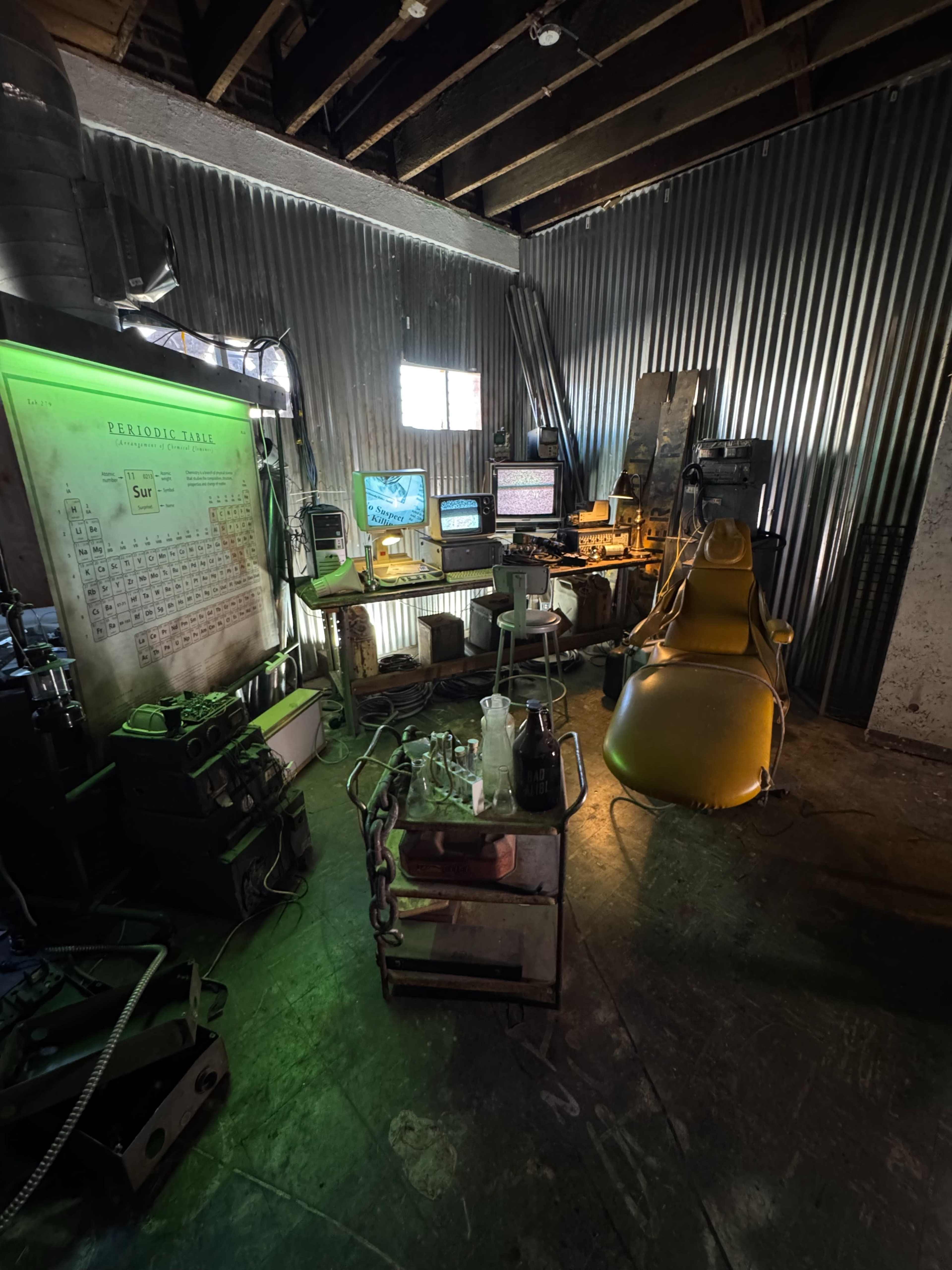 The image shows a dimly lit industrial workspace featuring a yellow chair, multiple computer screens, laboratory equipment, and a wall with a periodic table.