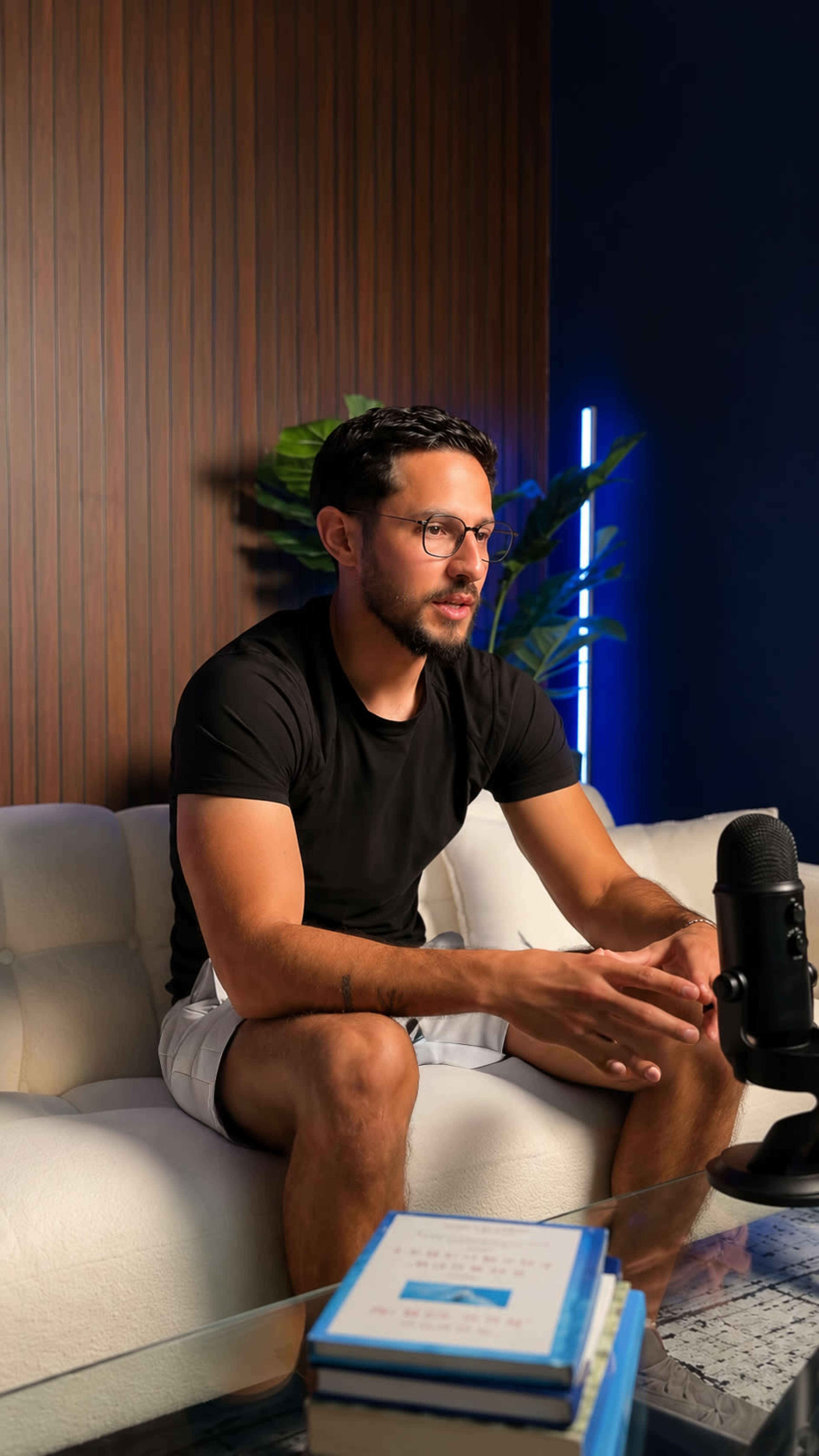 A man wearing a black t-shirt and shorts sits on a white sofa in a room with wooden paneling, speaking toward a microphone.