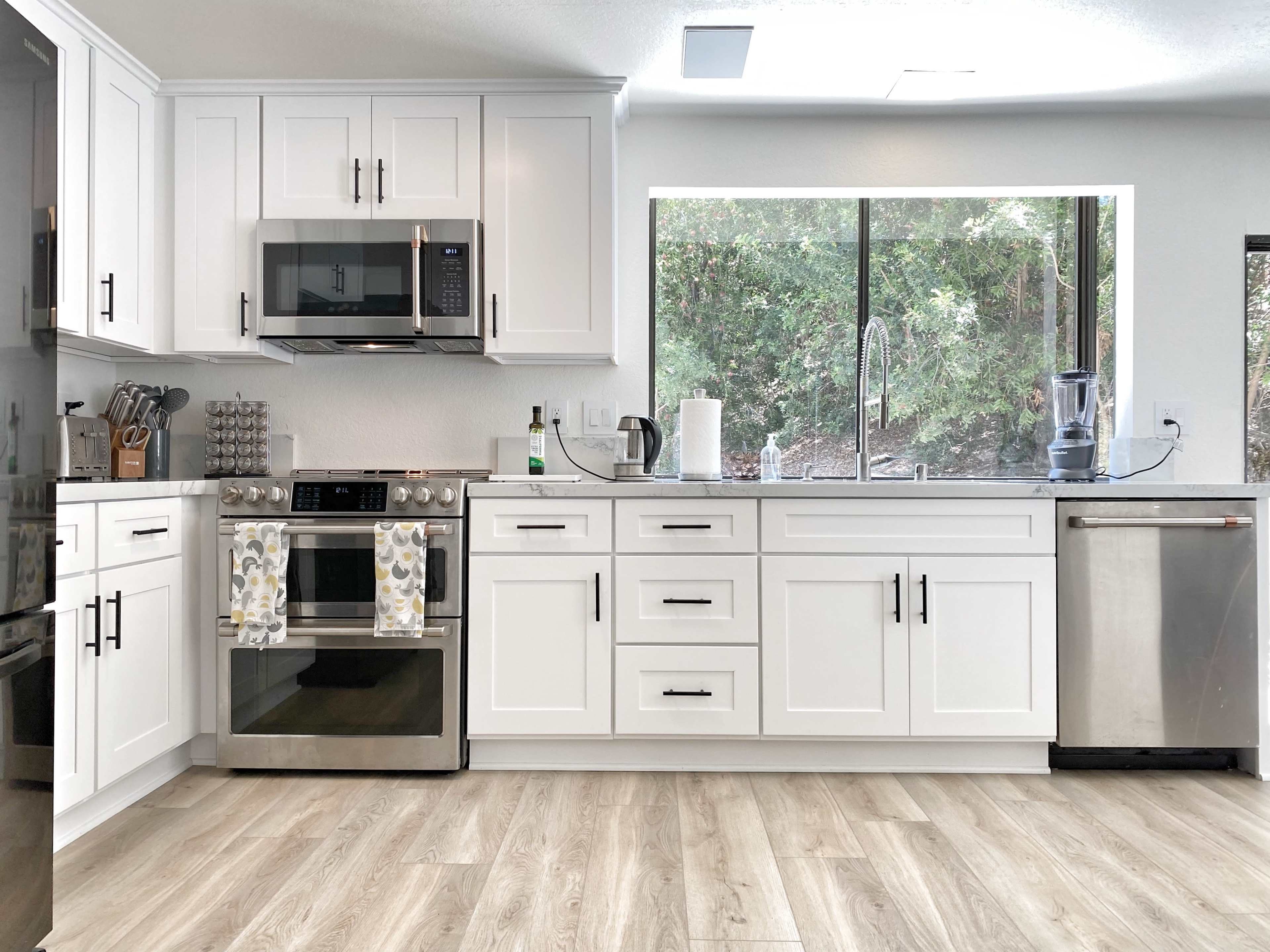 A modern kitchen features white cabinetry, stainless steel appliances, and a large window overlooking greenery.