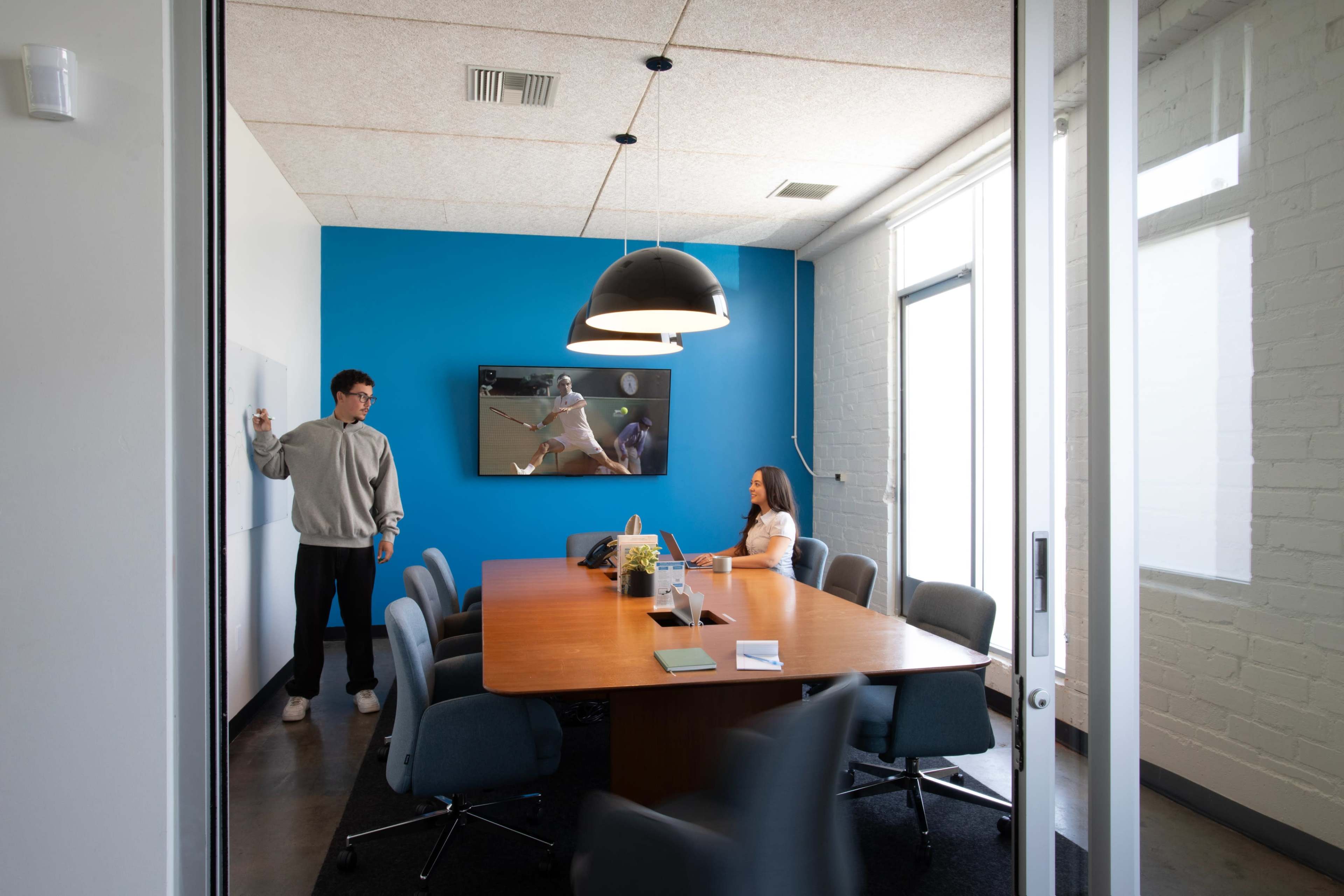 A person stands near a whiteboard while another sits at a conference table in a well-lit meeting room with a blue accent wall and a television displaying a sports event.