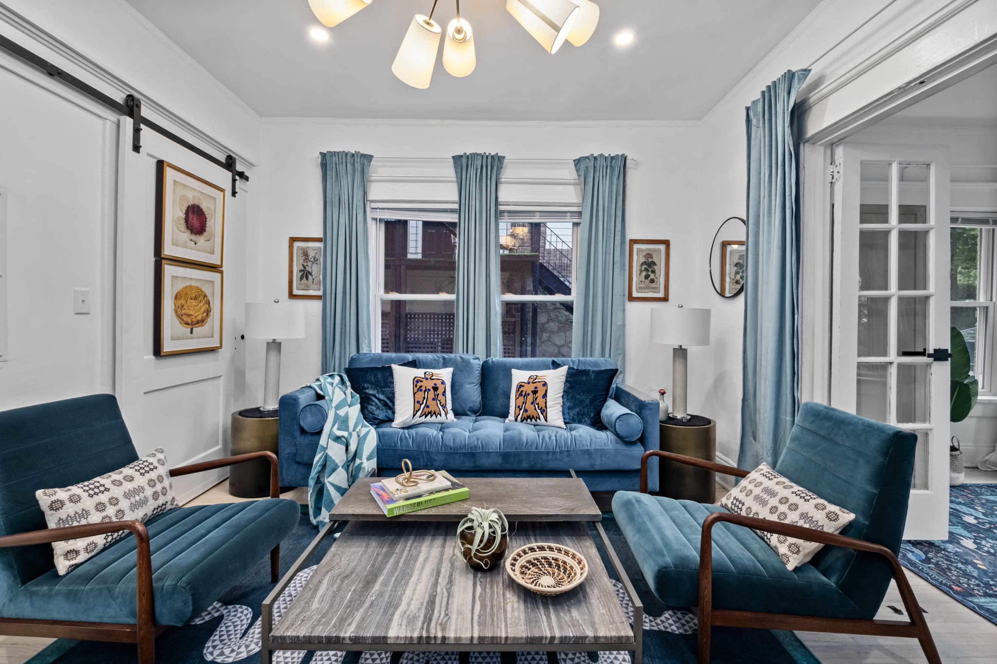 The image shows a cozy living room furnished with a blue sofa, two wooden chairs, and a coffee table, with large windows dressed in light blue curtains.
