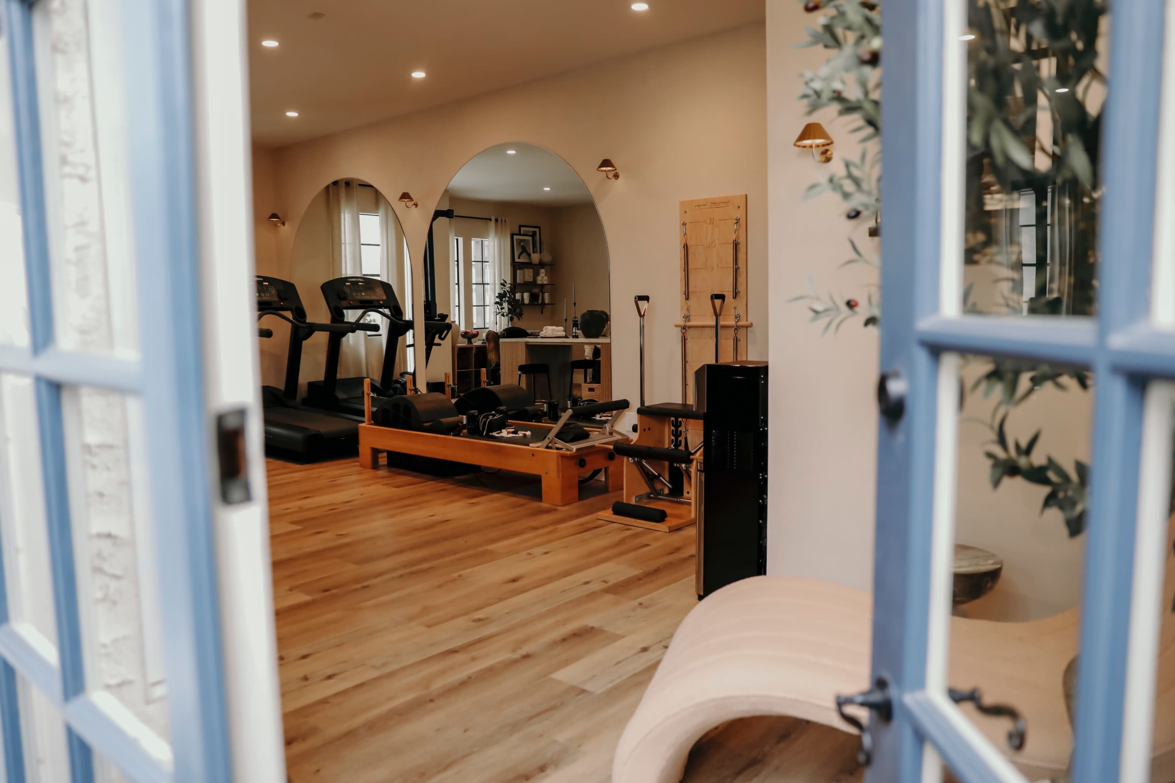 The image displays a home gym featuring treadmills, a rowing machine, and wooden flooring, framed by blue doors.