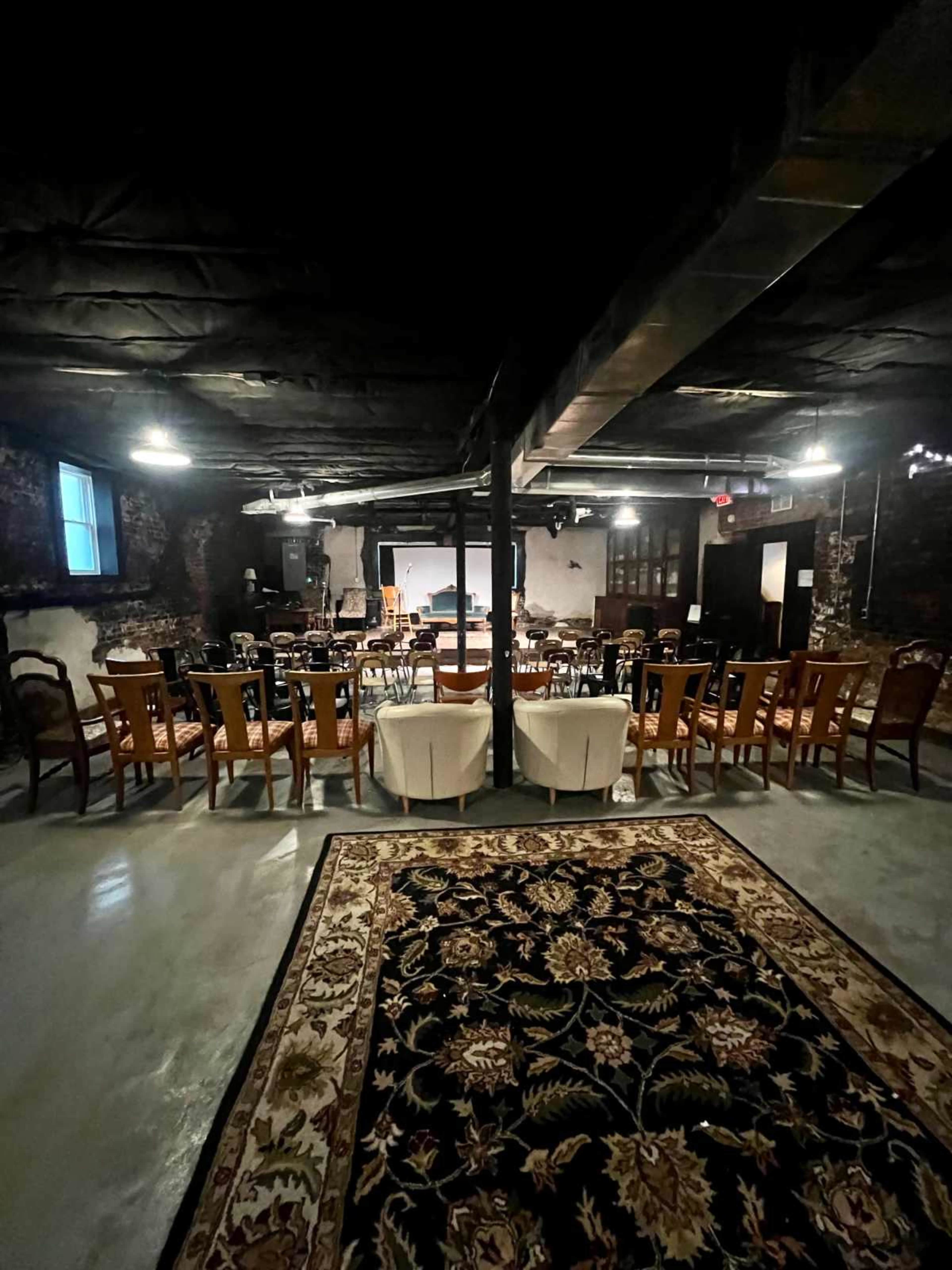 The image shows a dimly lit basement space set up for an event, featuring rows of chairs and tables arranged around a central stage area.