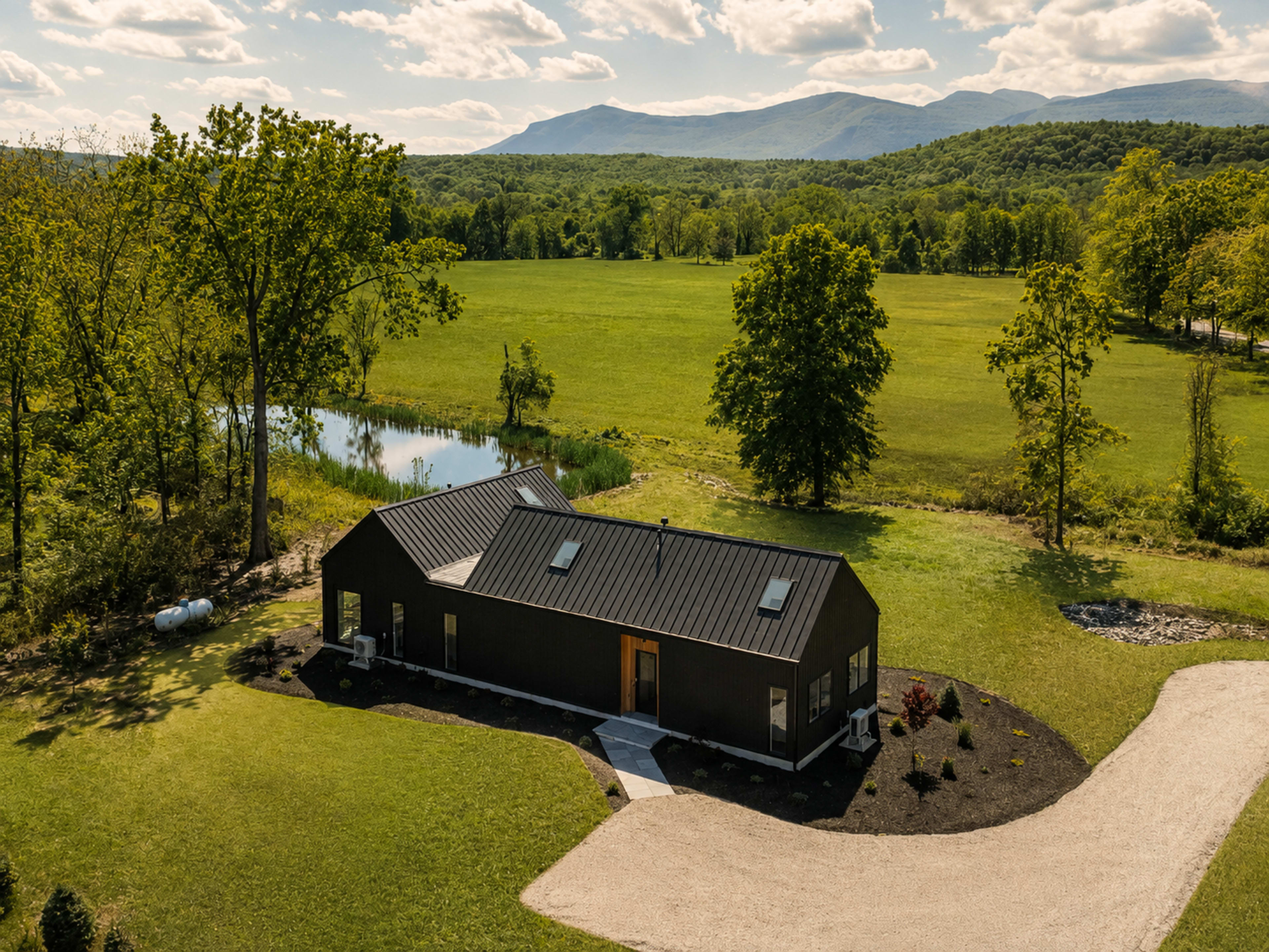 A modern black house is situated on a grassy landscape near a winding stream with mountains in the background.