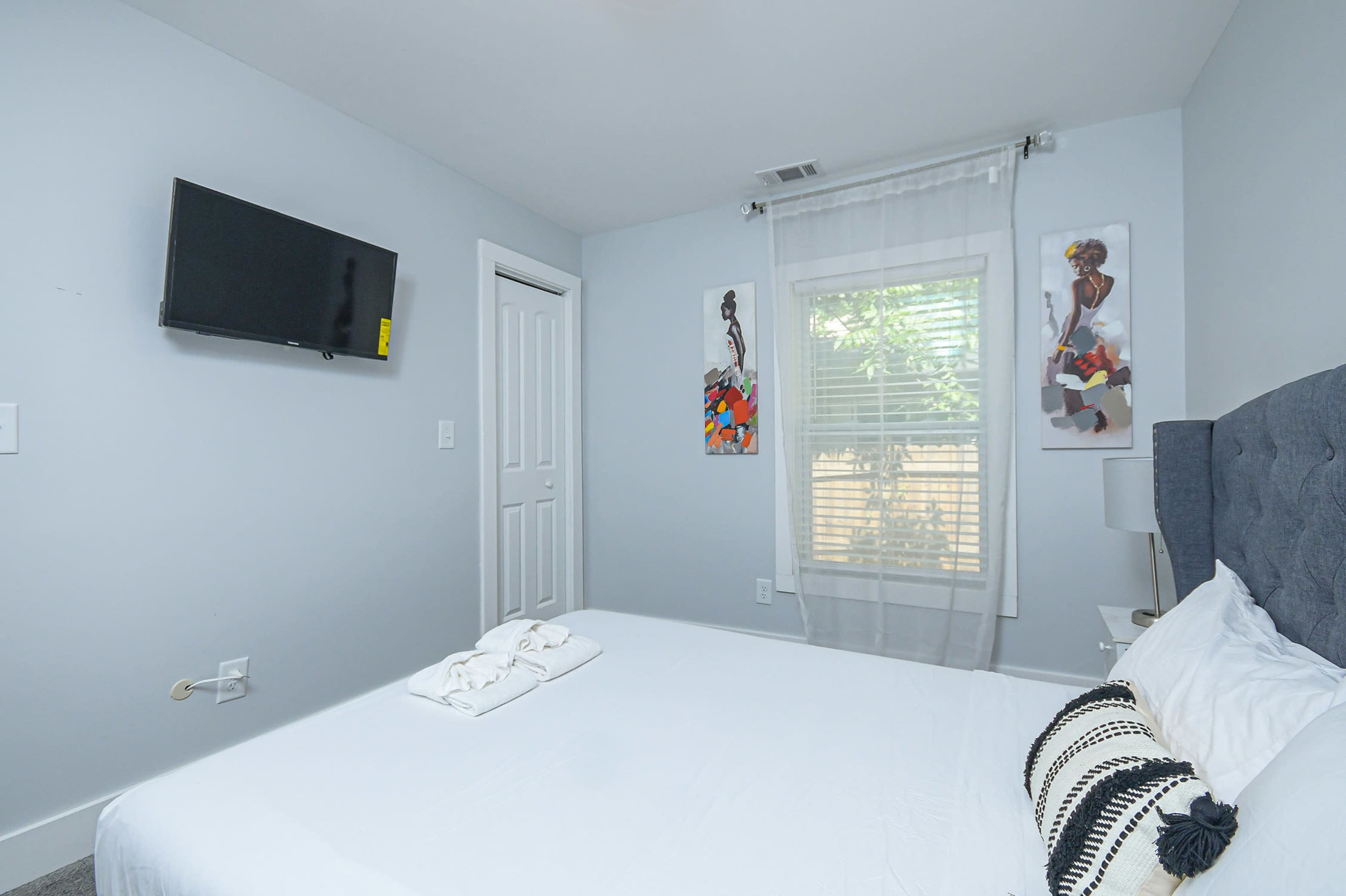 A bedroom features a bed with white bedding, a wall-mounted television, and two framed artworks beside a window with sheer curtains.