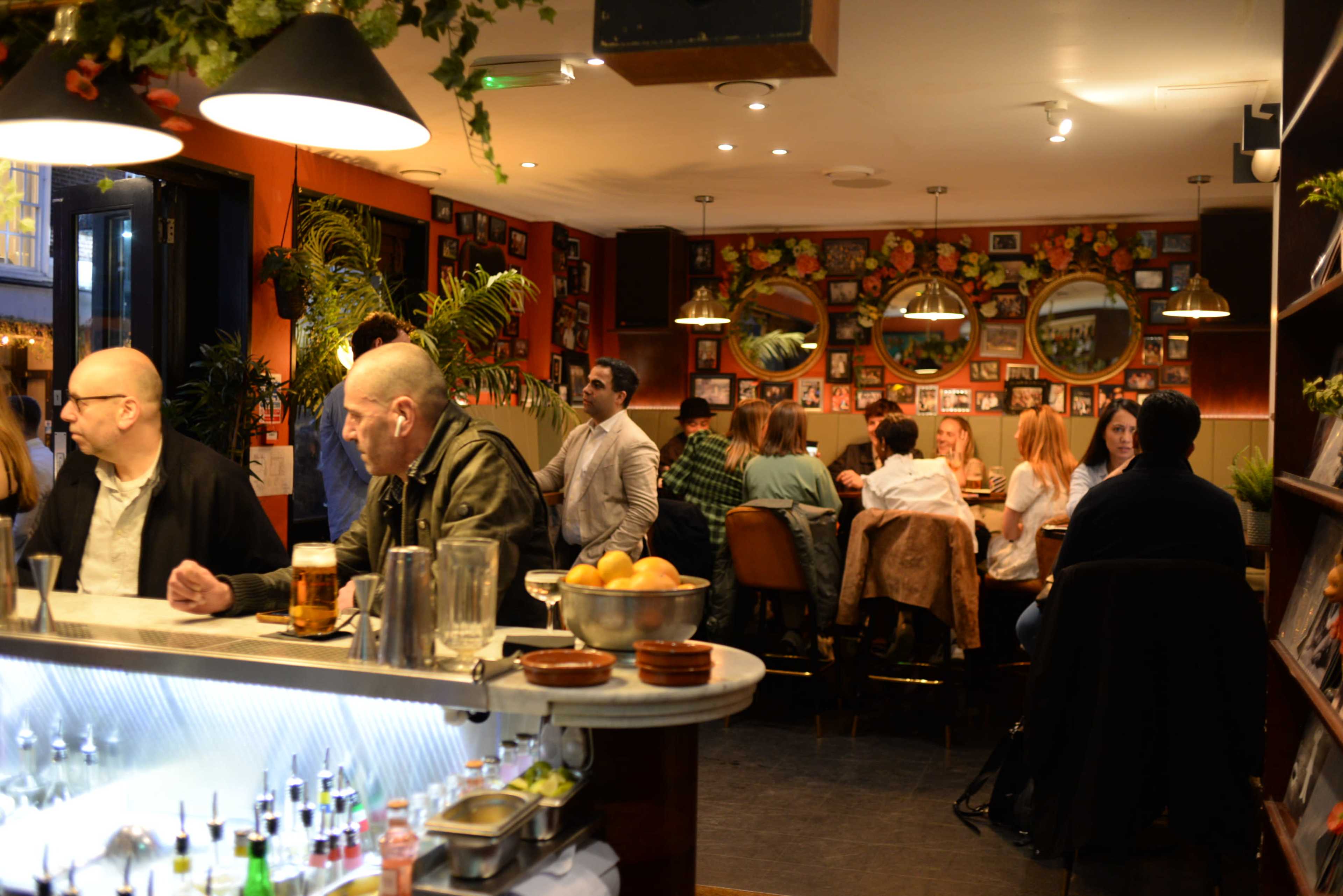 A bustling bar scene features patrons seated at tables and at the bar, surrounded by decorative plants and mirrors on the walls.