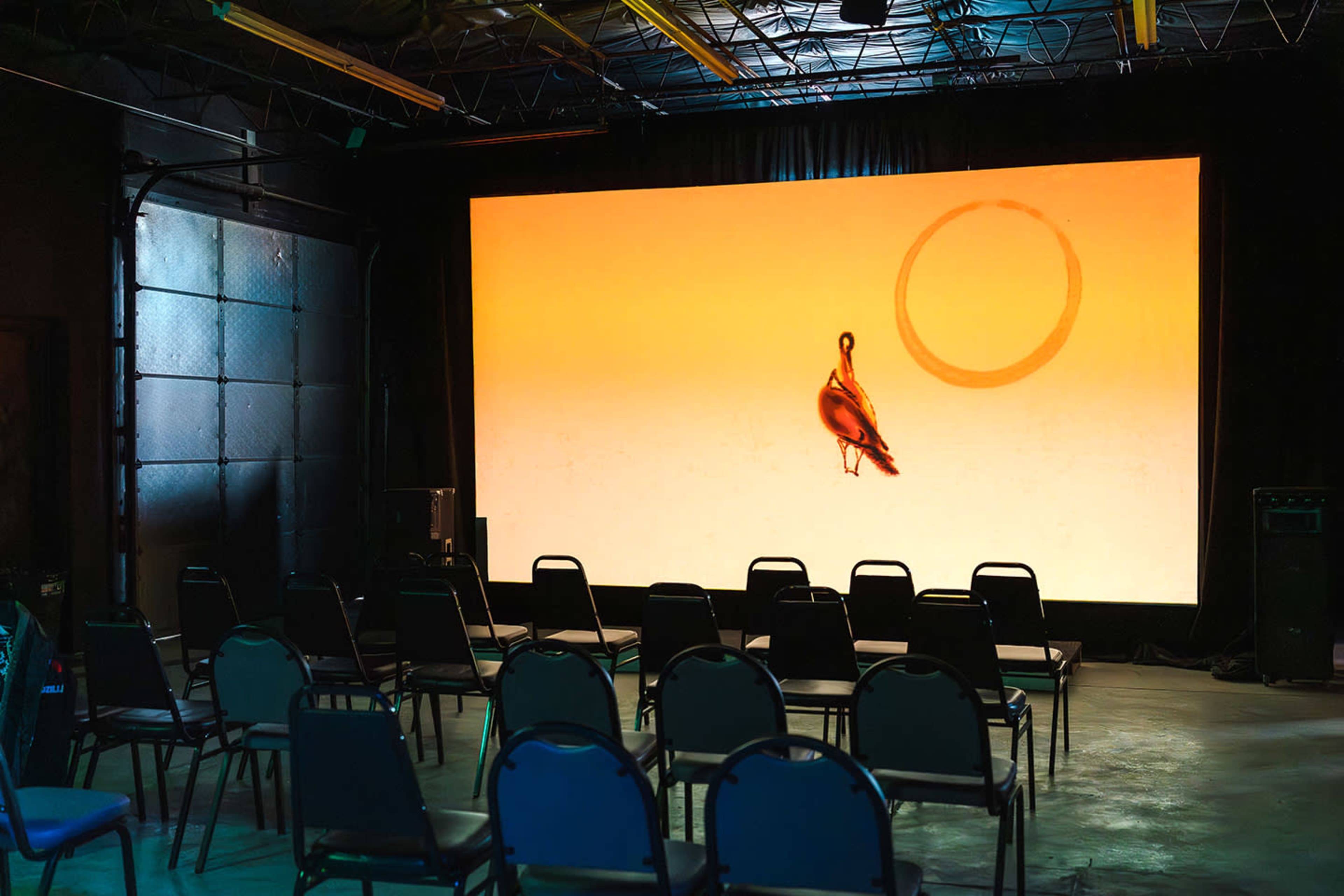 The image shows a projection screen displaying a bird against an orange backdrop, with a row of empty chairs arranged in front.
