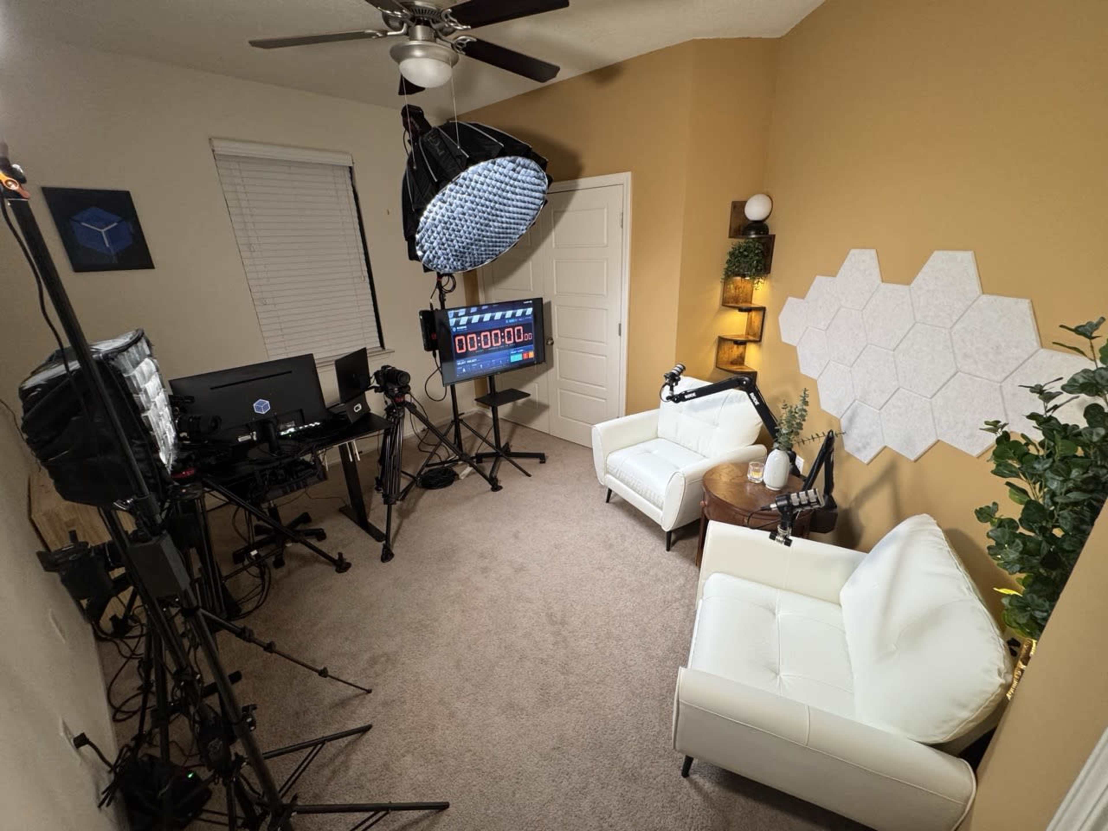 The image shows a small studio setup with two white armchairs, a circular table, and multiple cameras and lighting equipment arranged around a beige wall.