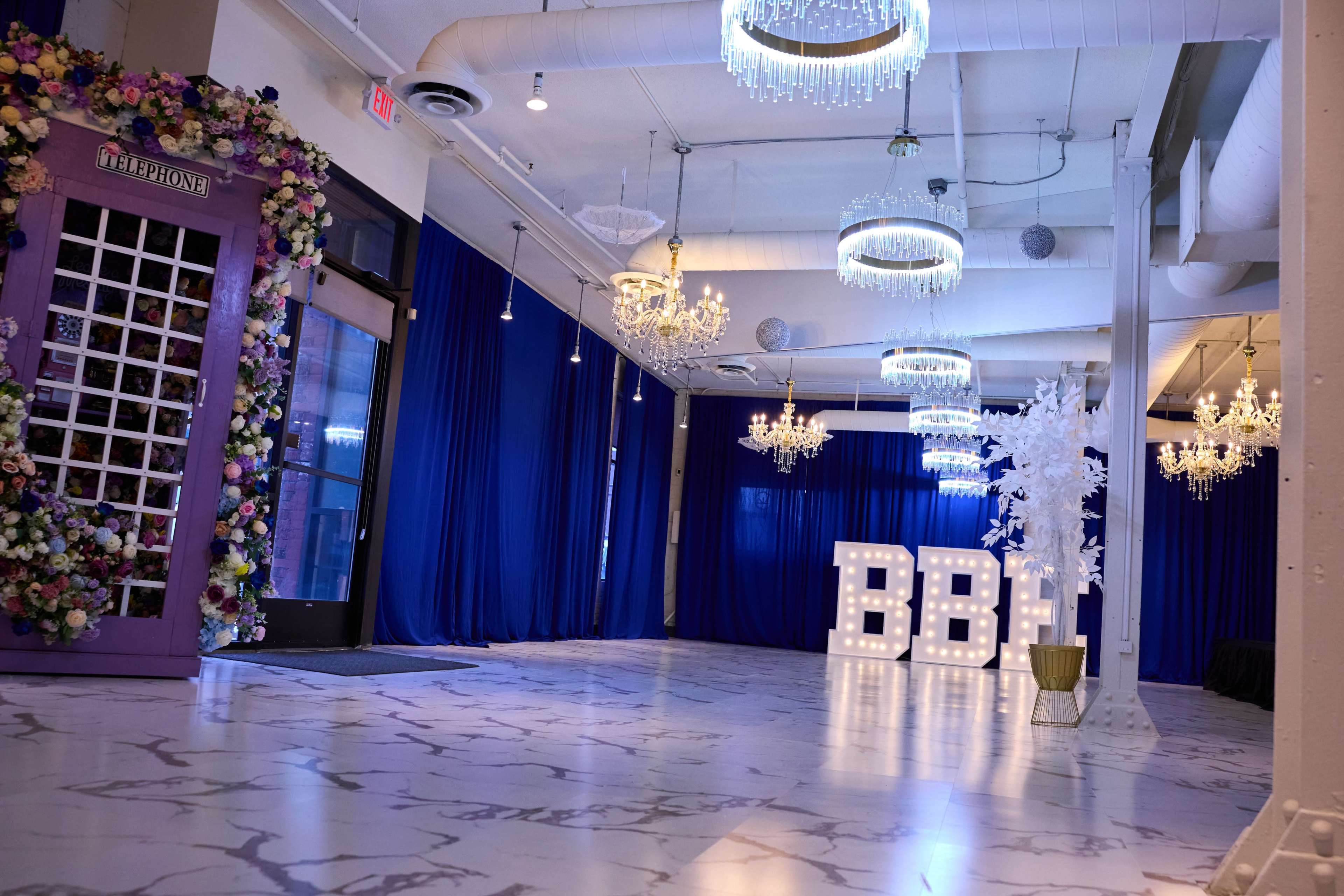 The image shows an elegantly decorated event space with a vintage telephone booth adorned with flowers, large illuminated letters spelling "BBI," and chandeliers hanging from the ceiling against a backdrop of blue curtains.