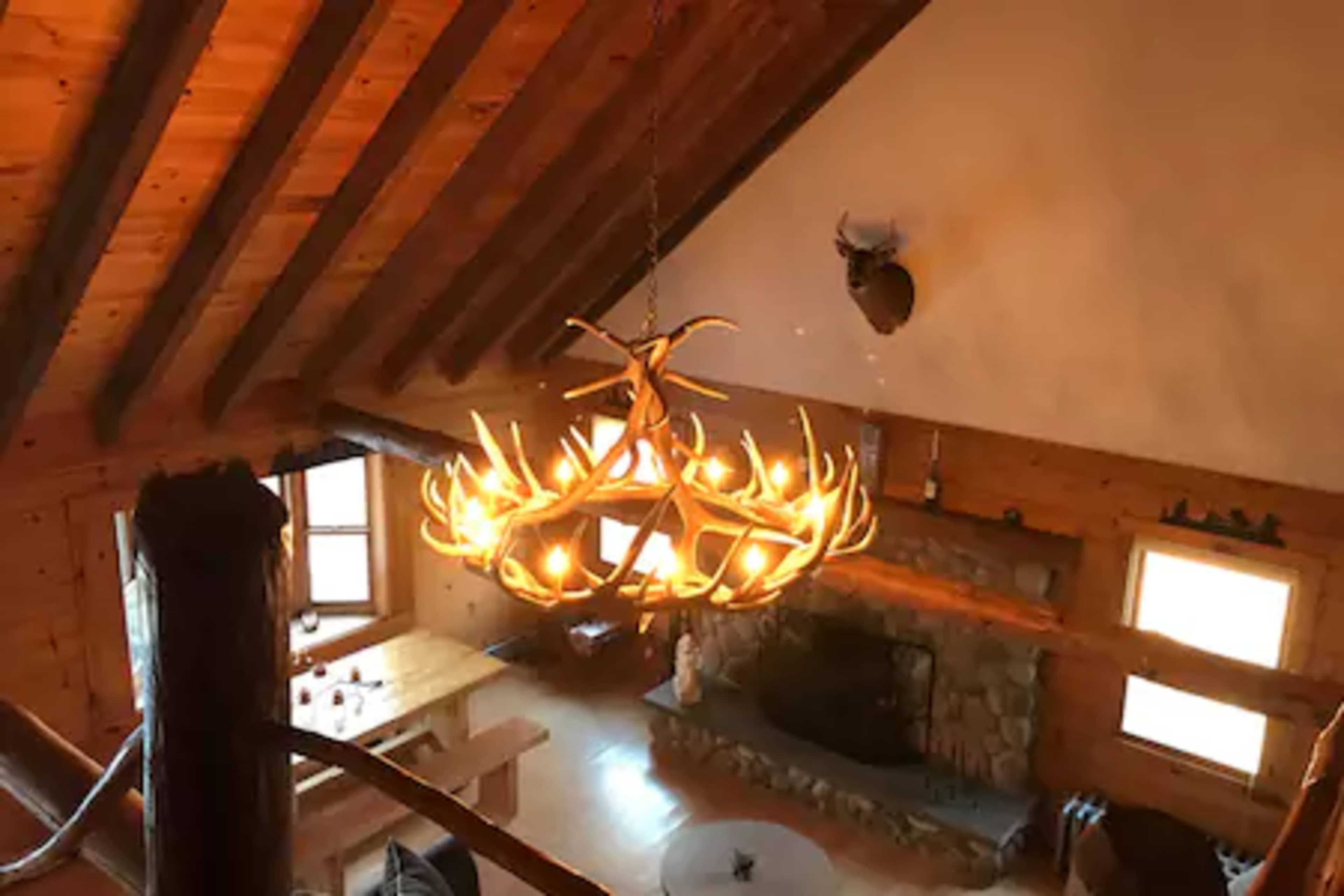 A rustic wooden living space with a large antler chandelier, a stone fireplace, and a dining table.