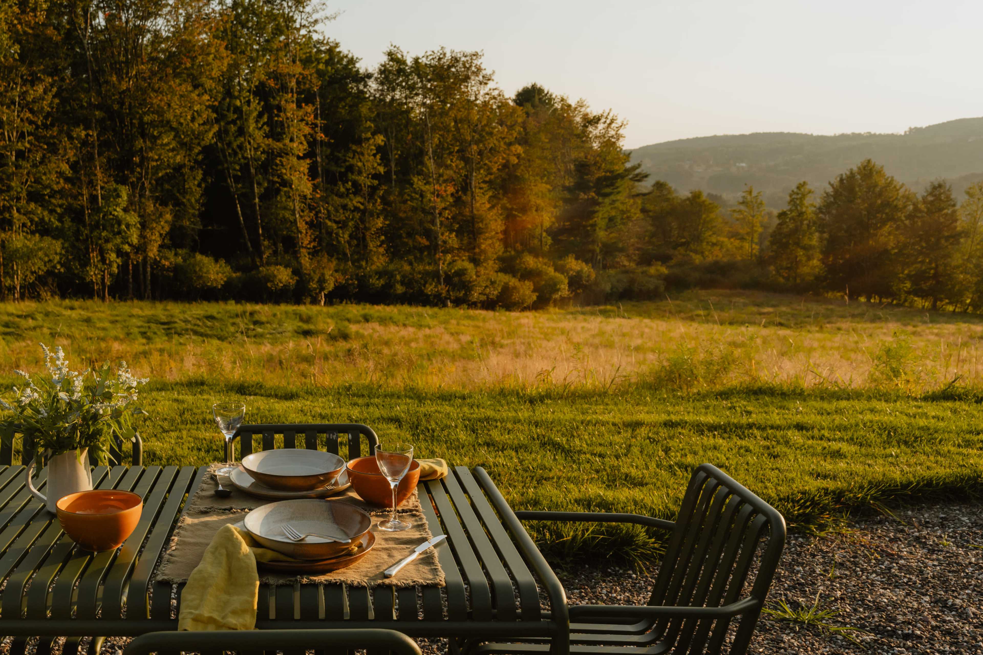 A dining table is set outdoors with plates and bowls, overlooking a grassy field and distant trees.