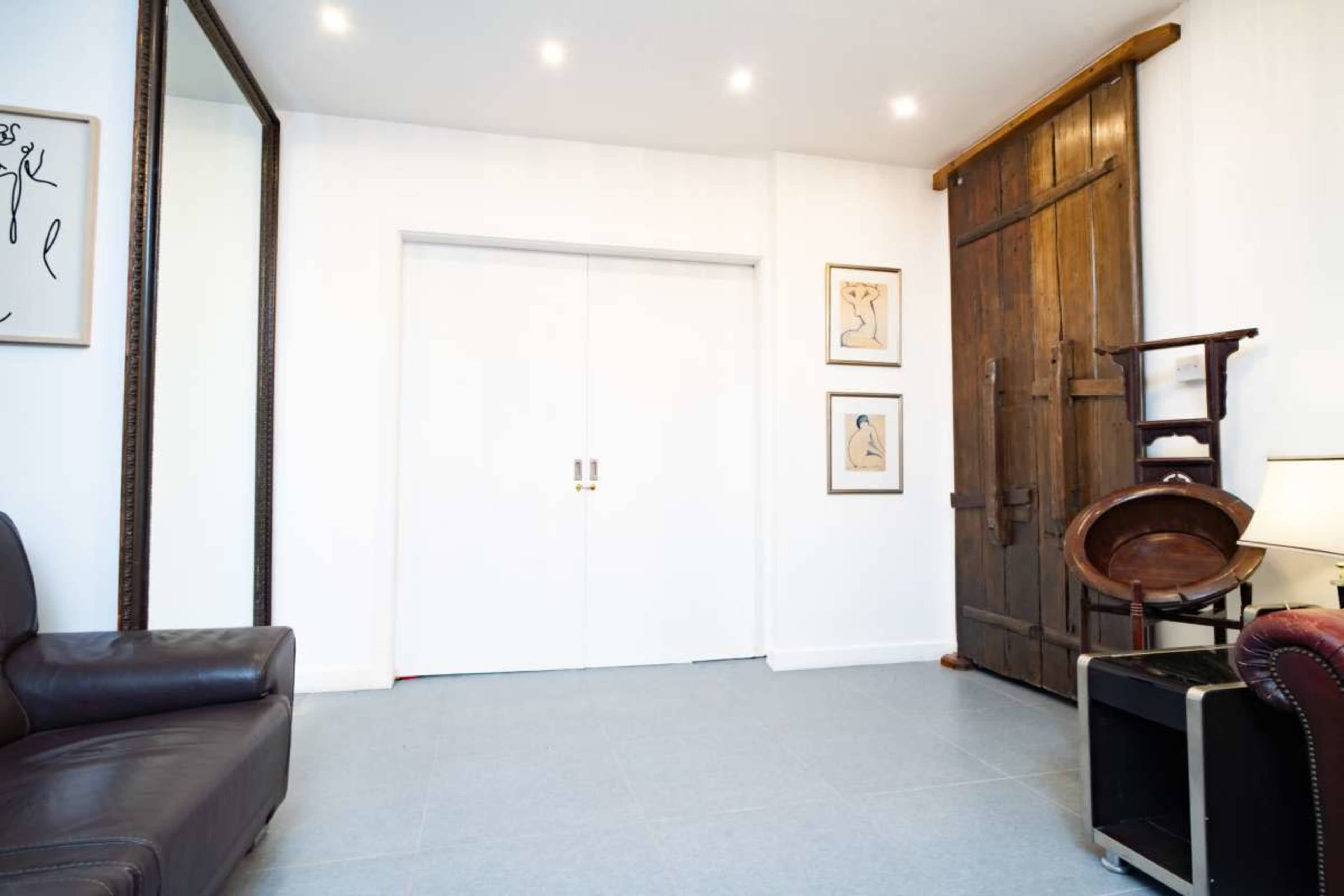 The image shows a modern interior space with a leather sofa, a large mirror, and a wooden door, complemented by framed artwork on the walls.