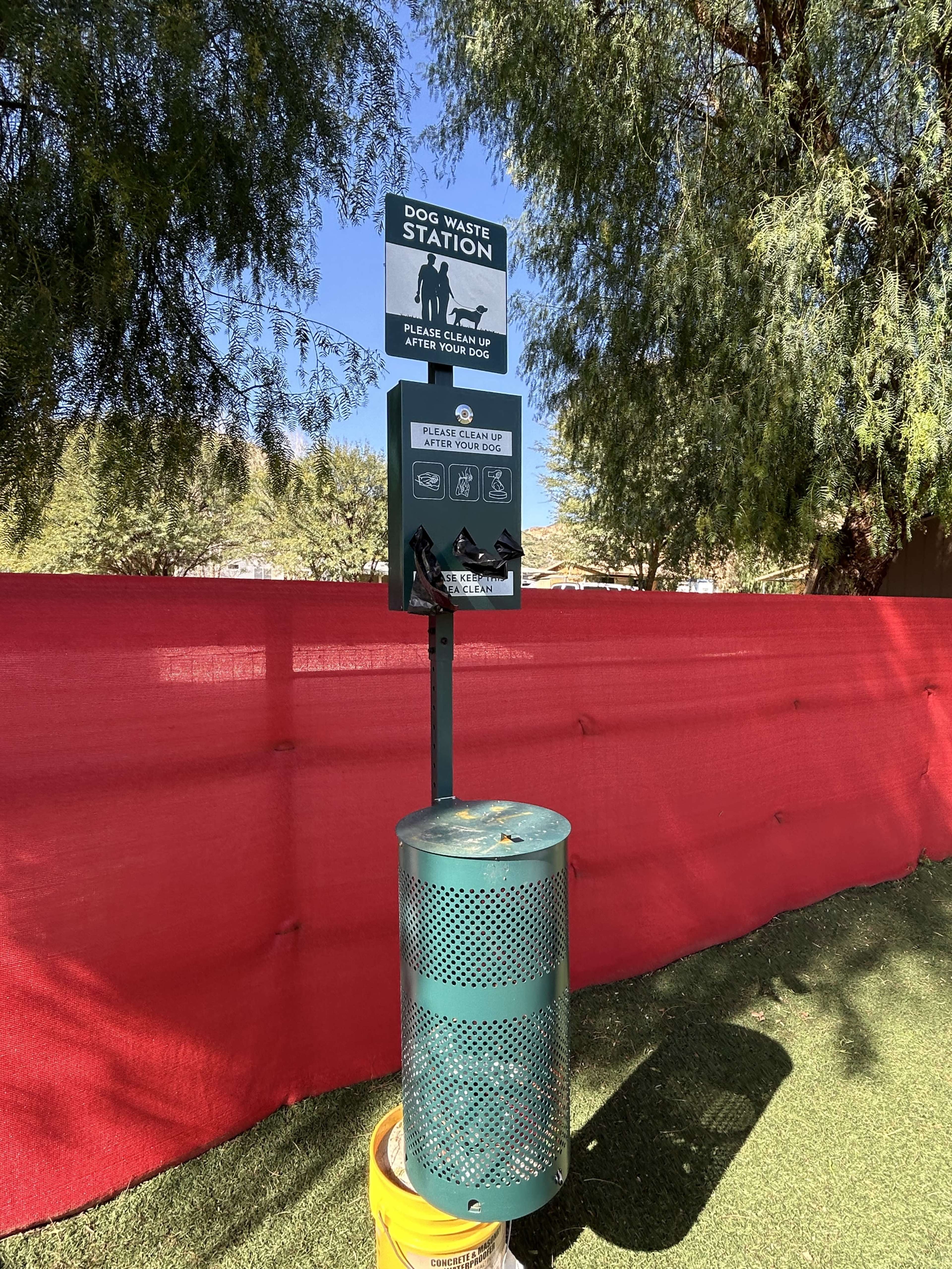 A dog waste station is installed in a park, featuring a sign with instructions and a container for disposing of pet waste.