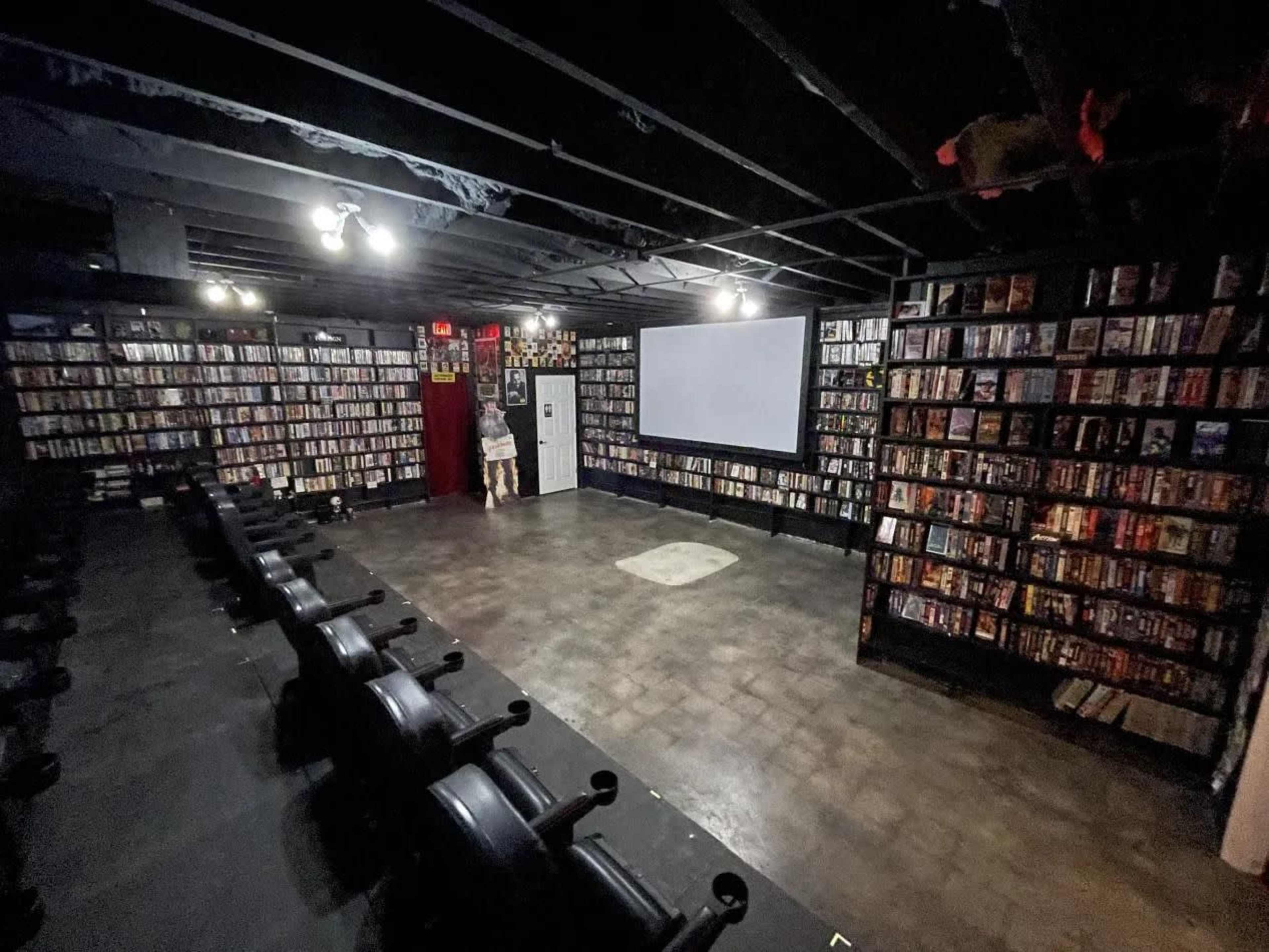The image shows a dimly lit, large room with shelves filled with DVDs lining the walls and a blank screen at the front.