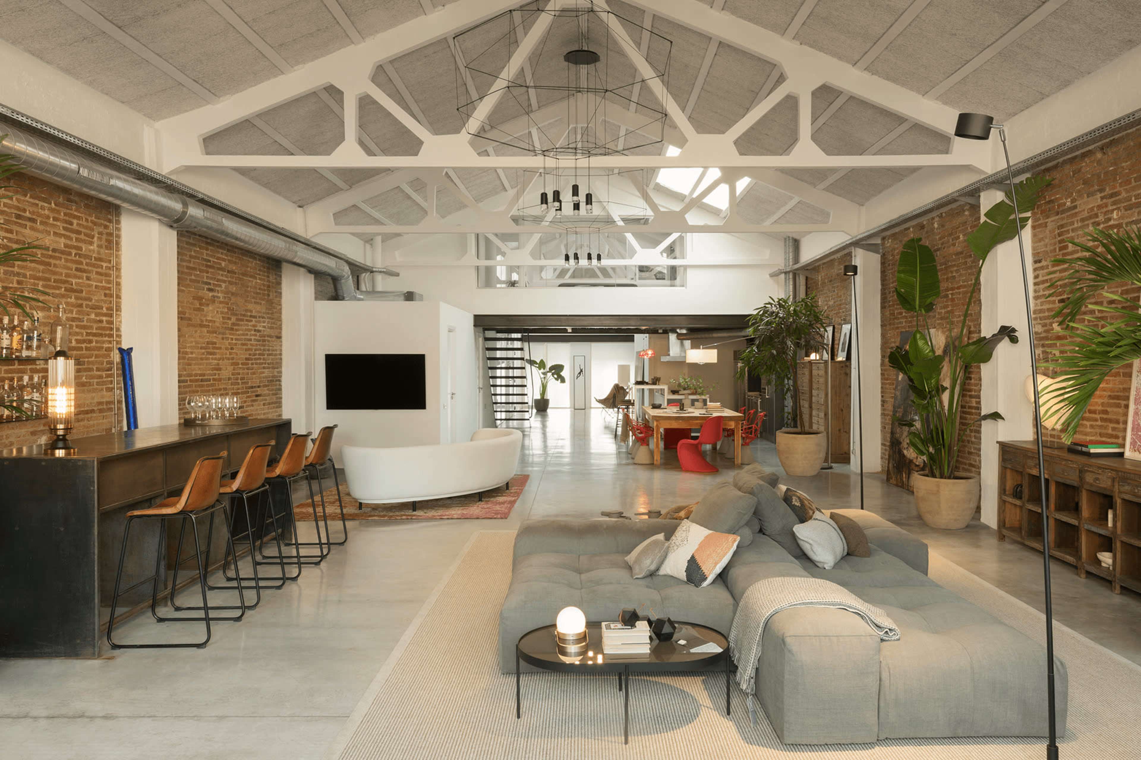 The open-concept living space features a large sofa, a bar area with high stools, and a dining area, illuminated by natural light through ample windows and complemented by decorative plants.