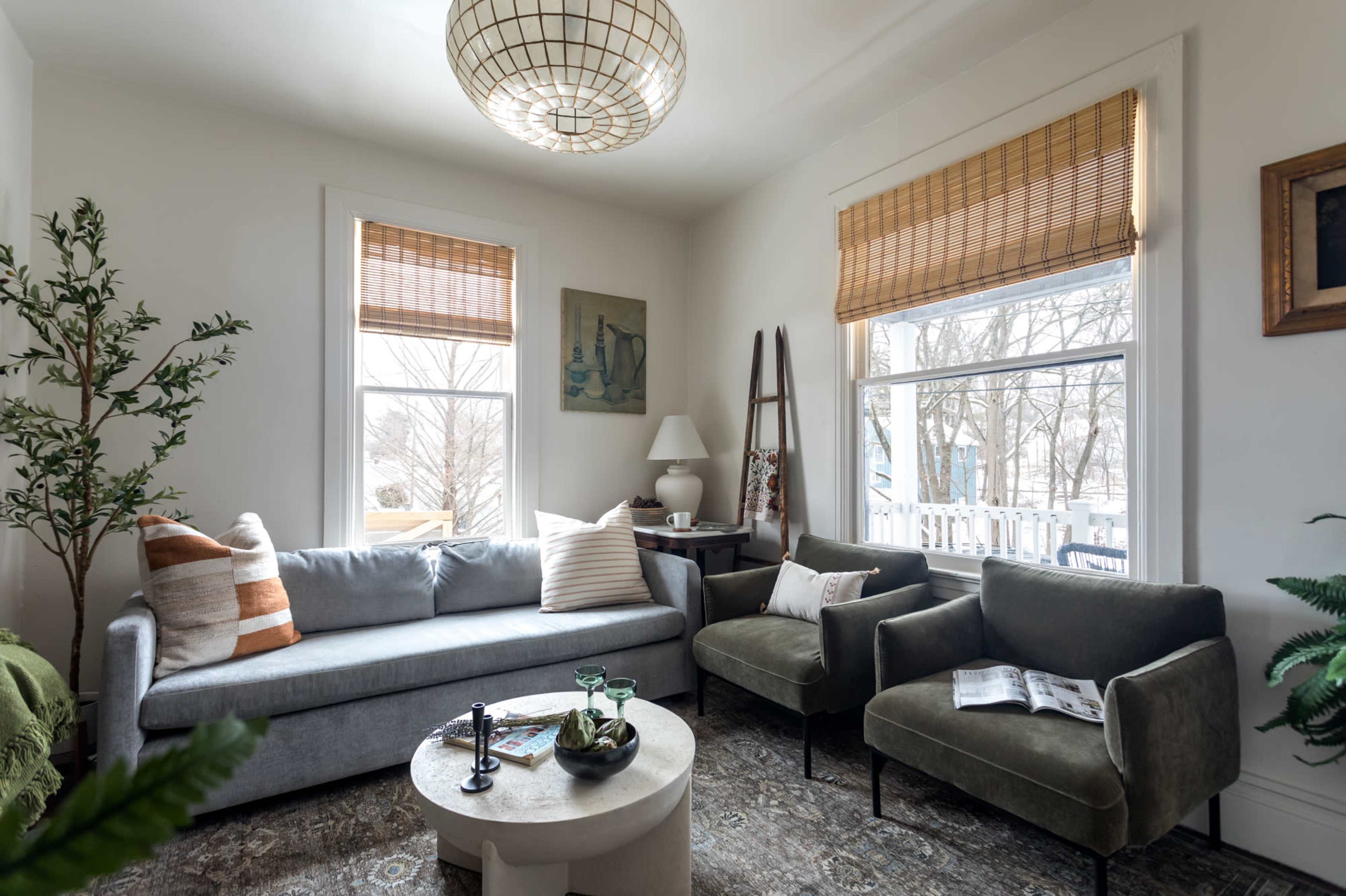 A cozy living room features a gray sofa, two green chairs, a round coffee table, and large windows with bamboo shades, surrounded by decorative plants.