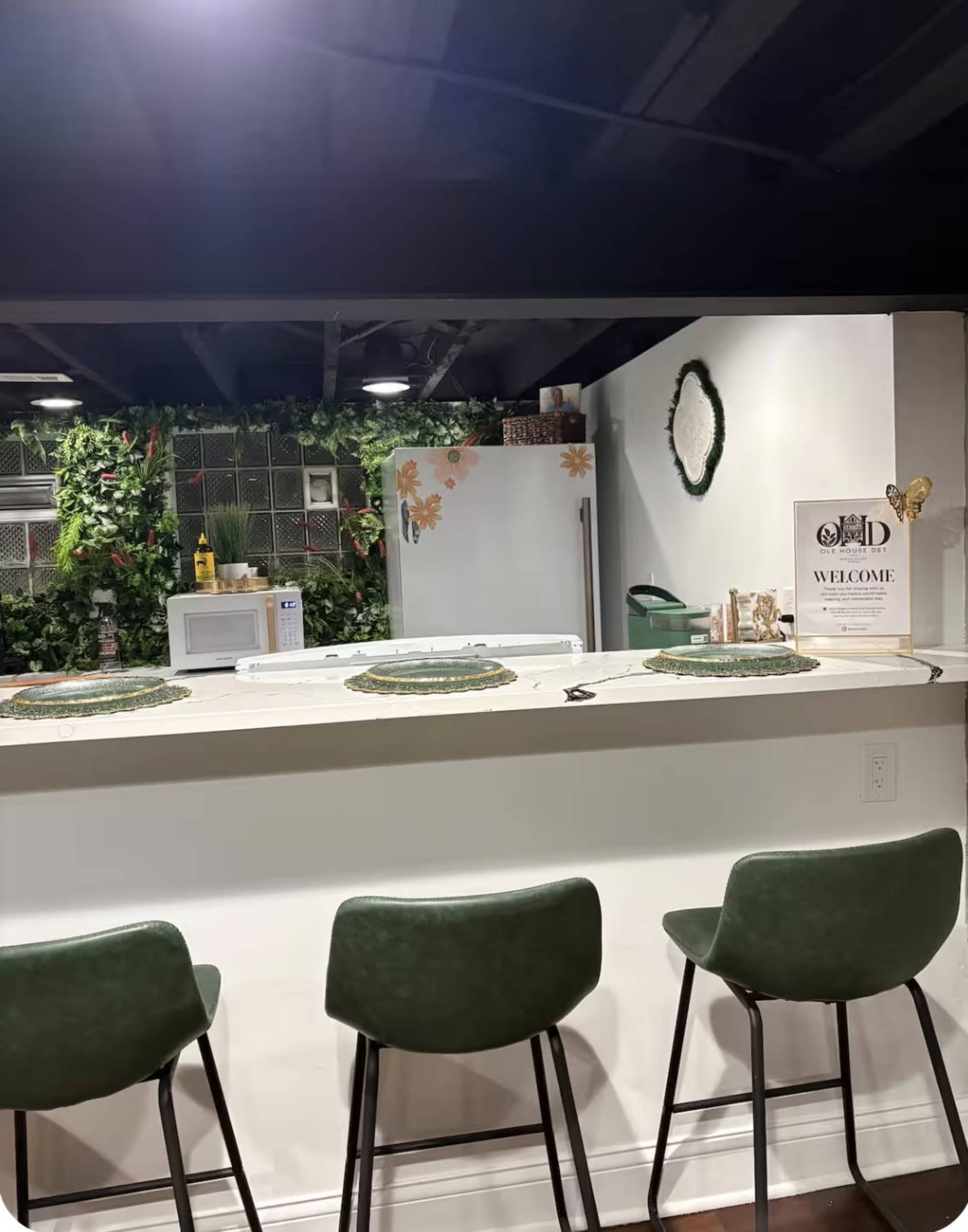 The image shows a modern kitchen bar area with green seating, a refrigerator, and decorative plants in the background.