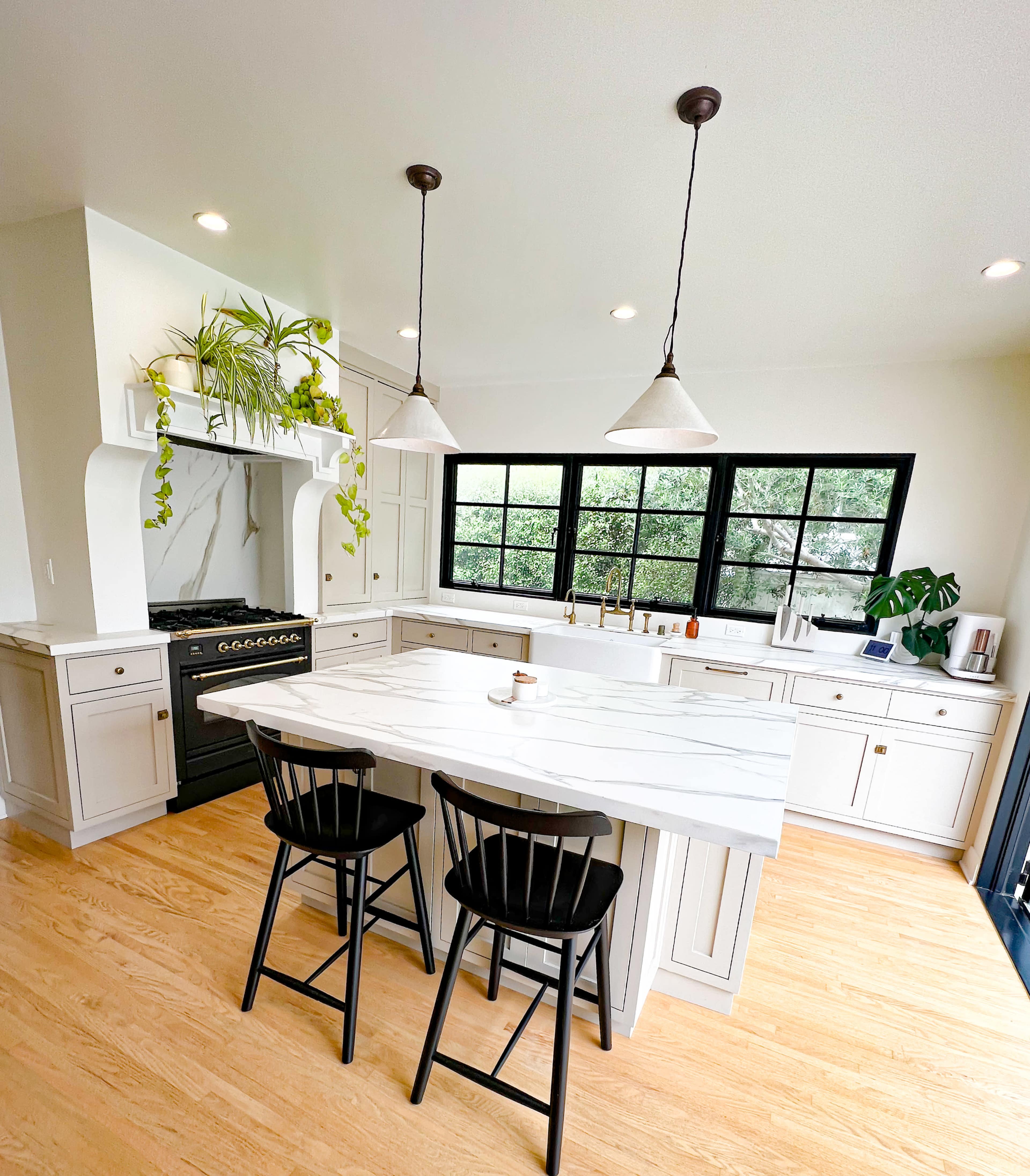 A modern kitchen features a marble island with two black stools, white cabinetry, large windows, and pendant lighting.