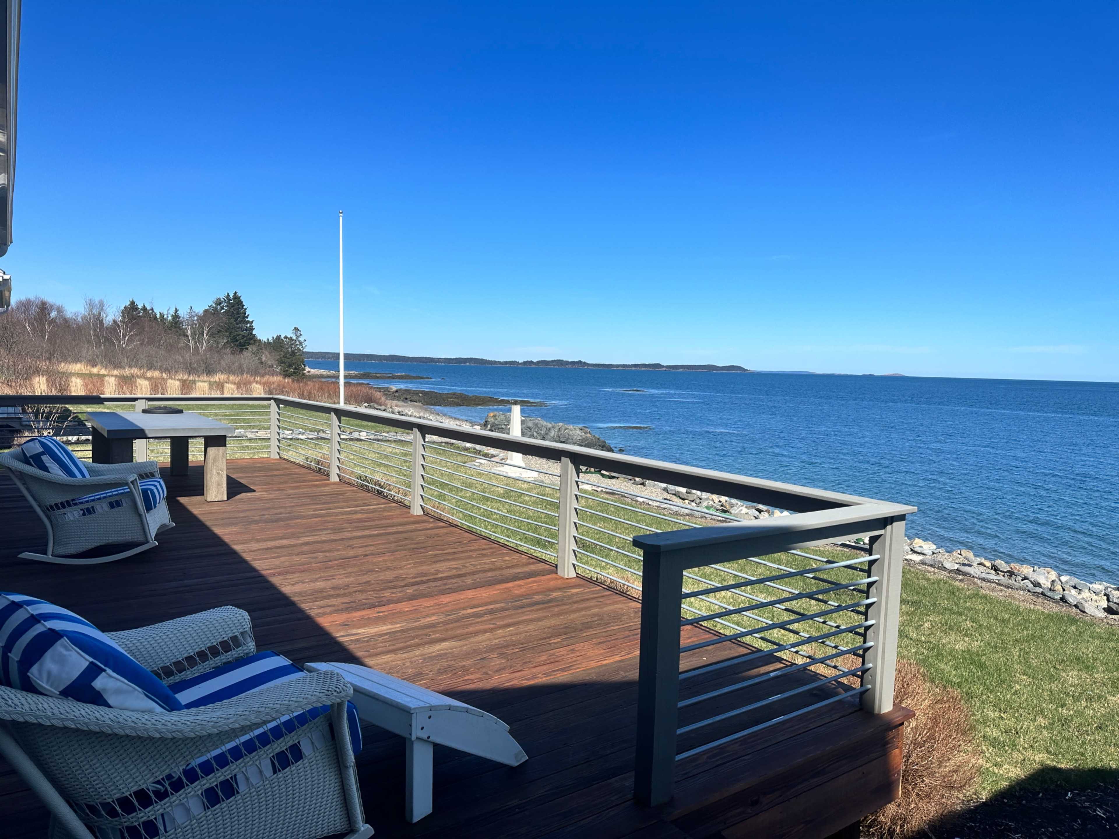 A wooden deck with white and blue-striped chairs overlooks a calm shoreline and clear blue water.