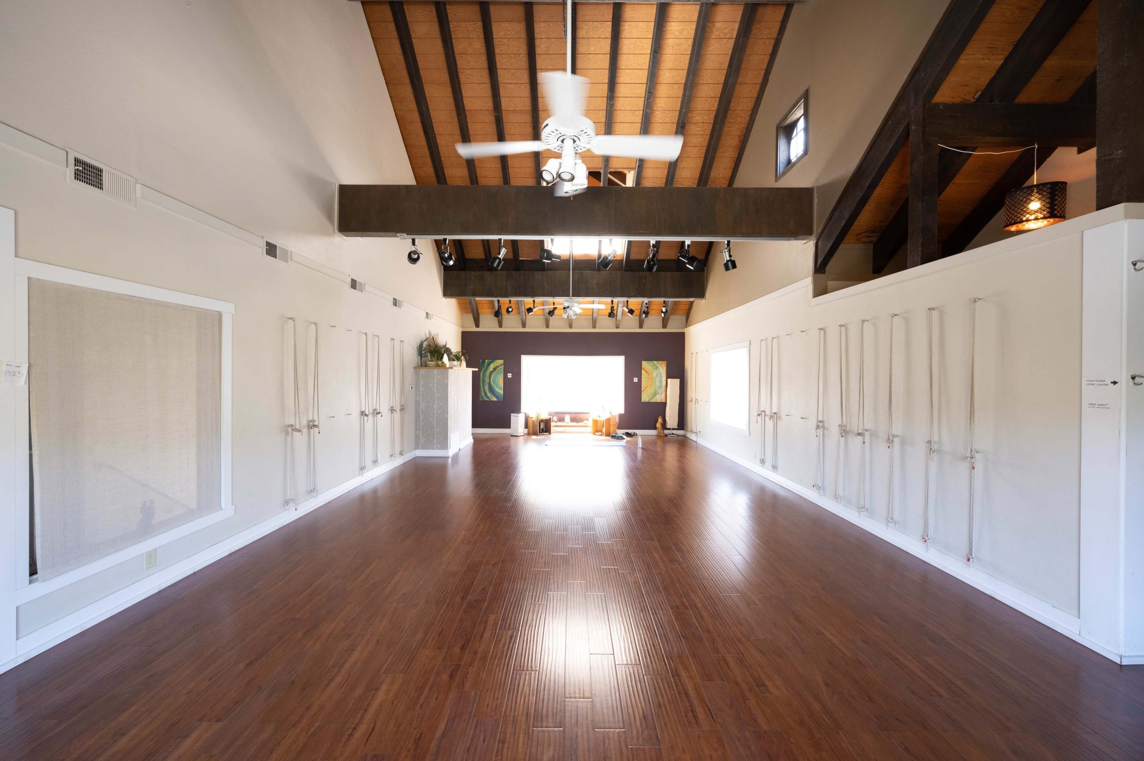 The image shows a spacious, well-lit yoga studio with wooden flooring and a high ceiling featuring exposed beams.