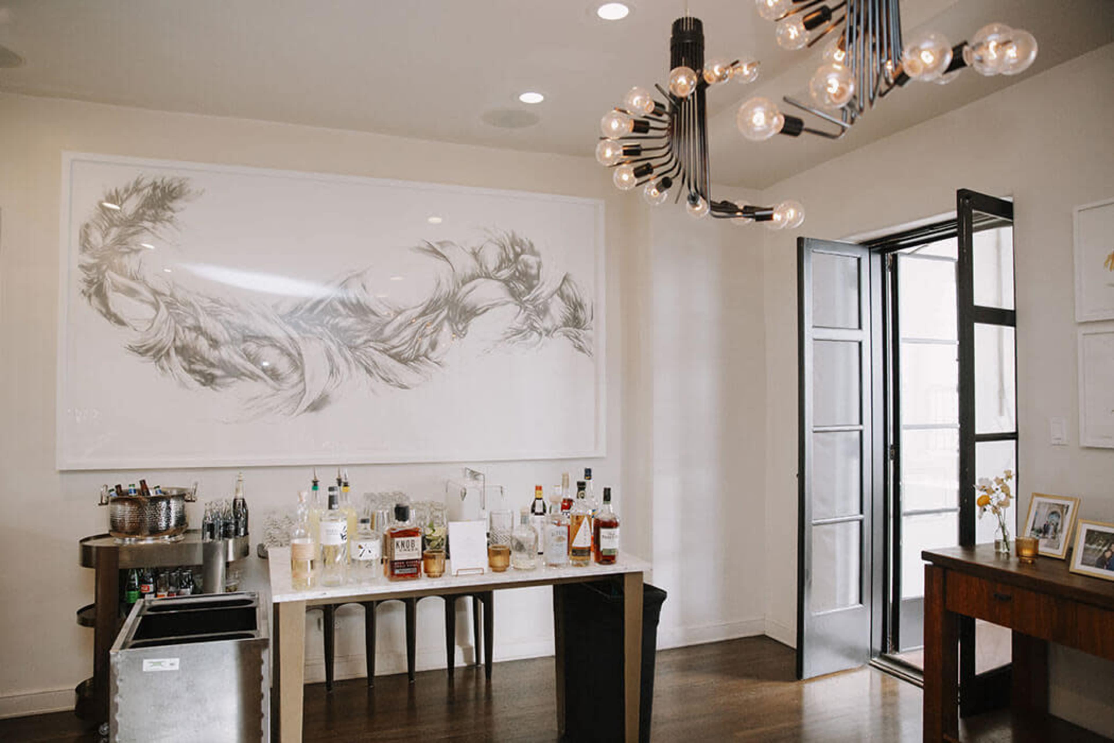 The image shows a modern interior with a bar area featuring various bottles of alcohol and an artistic wall piece above.