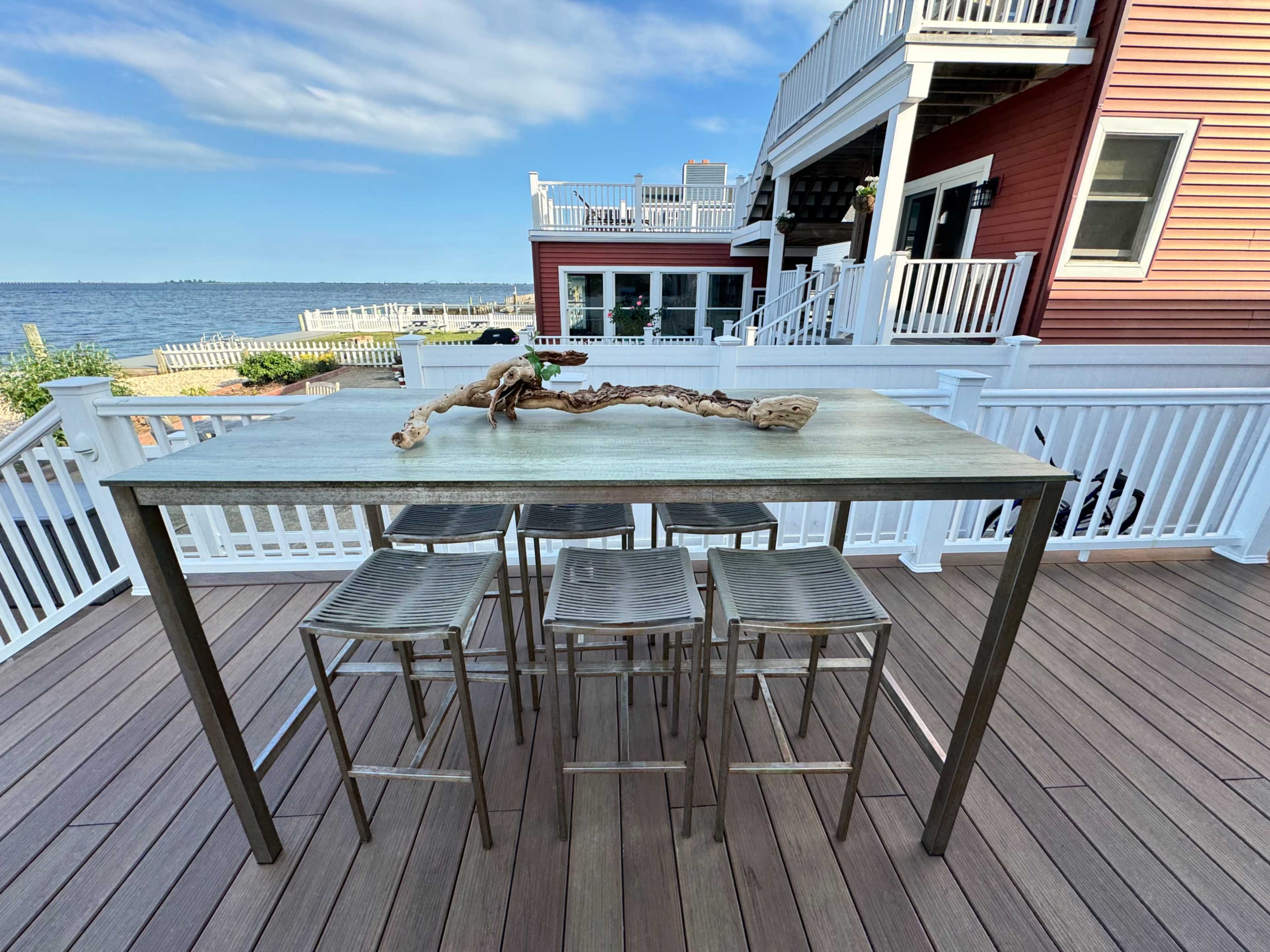 A modern outdoor bar table with metal stools overlooks a body of water and is adjacent to a waterfront house.