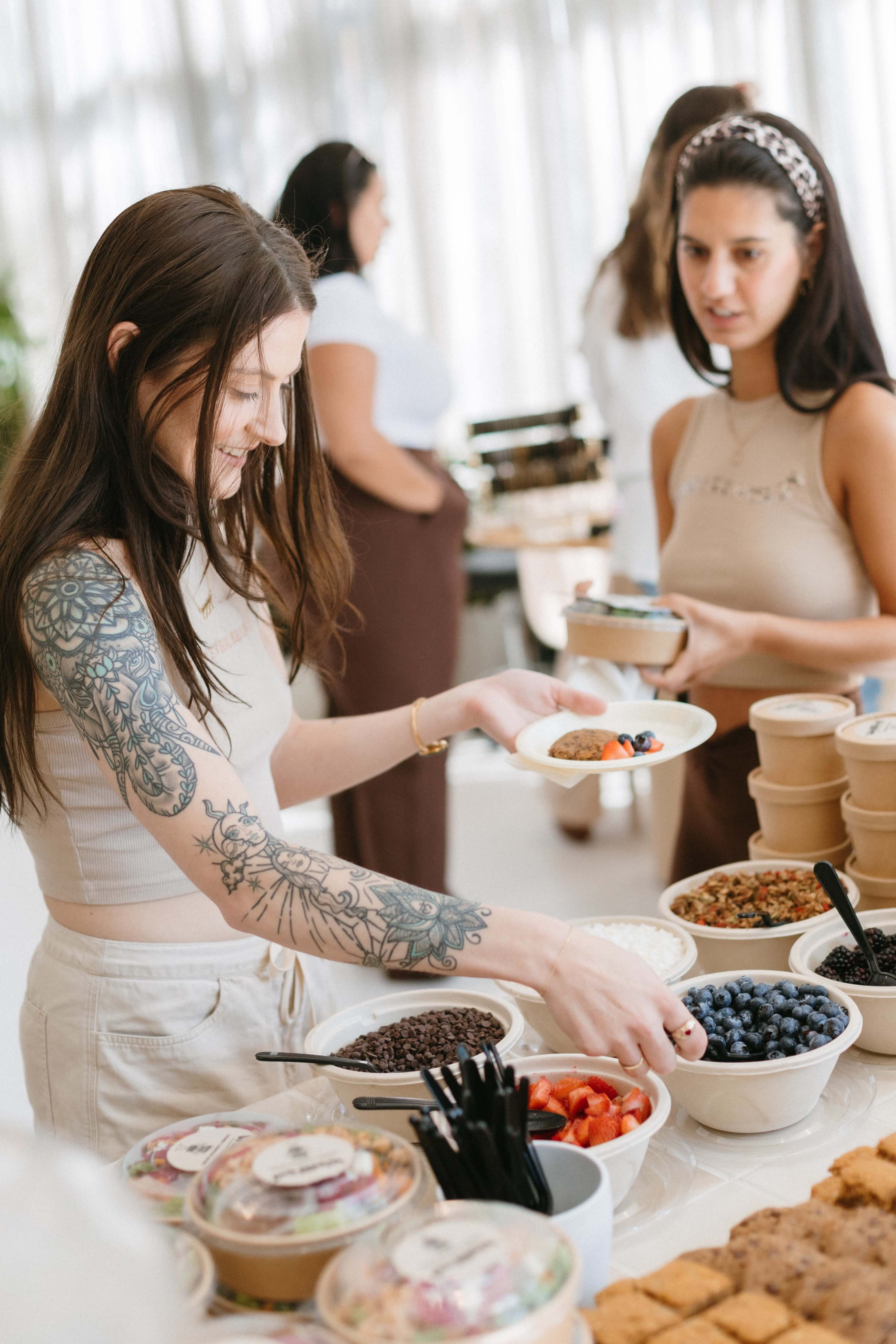 A woman with tattoos selects food from a buffet while another woman stands nearby, holding a phone.