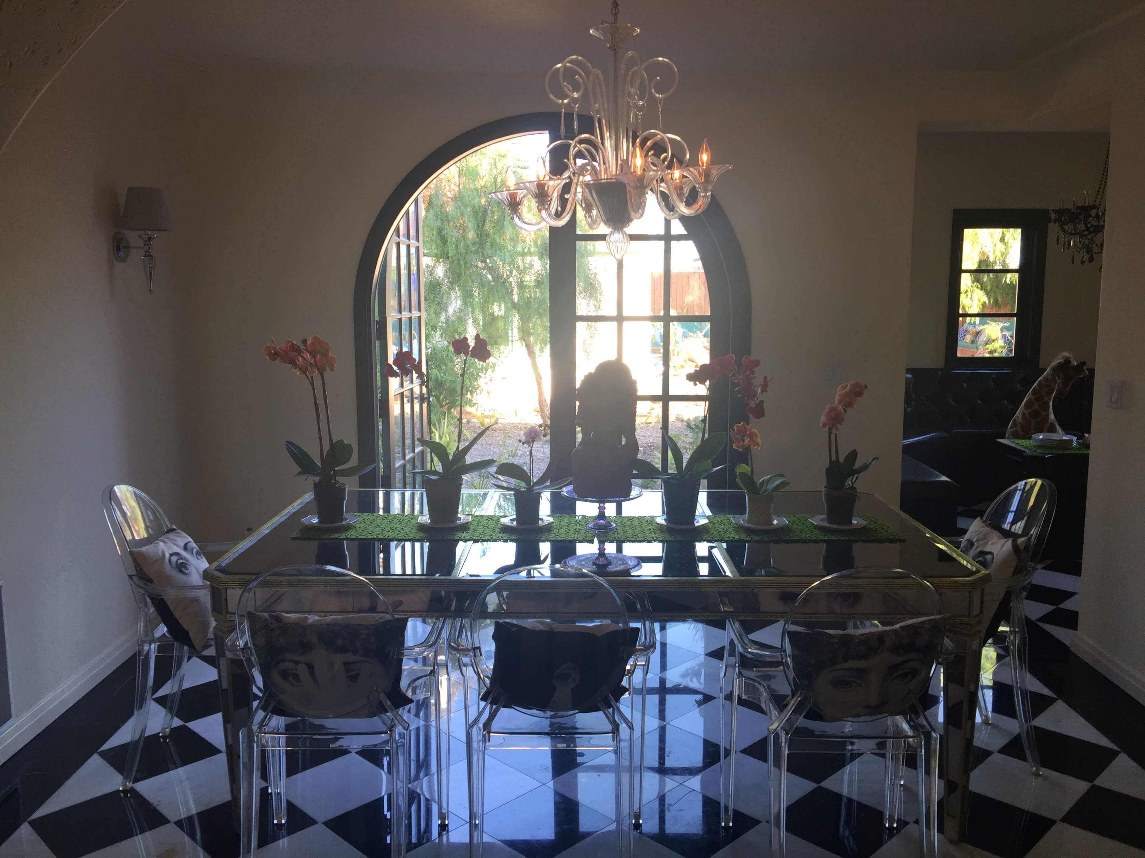 The image shows a dining room with a glass table surrounded by clear chairs, featuring potted orchids as a centerpiece, and large windows letting in natural light.