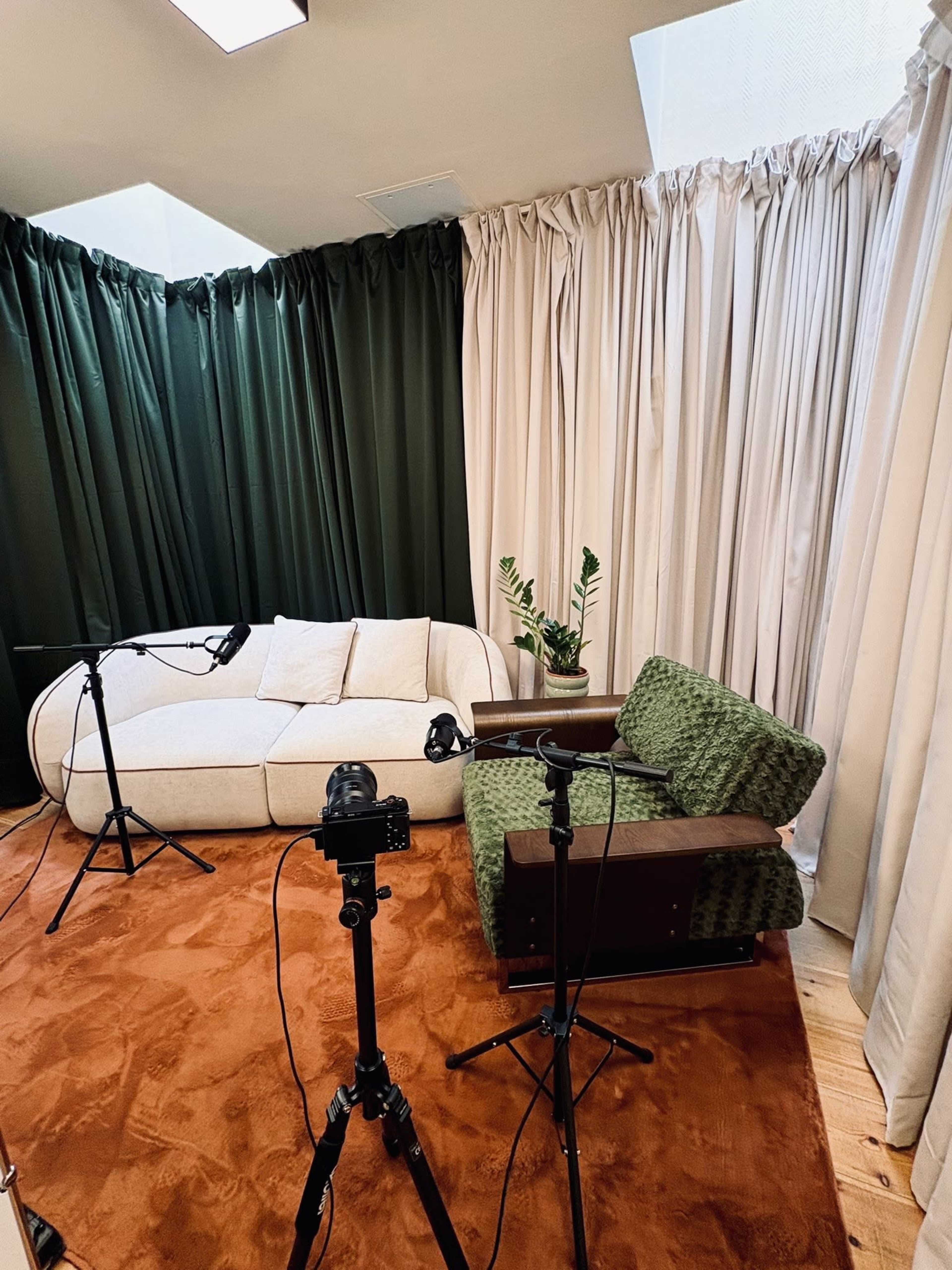 The image shows a cozy interior with a white couch and a green armchair on a brown rug, surrounded by tall curtains and equipped with camera tripods and microphones.