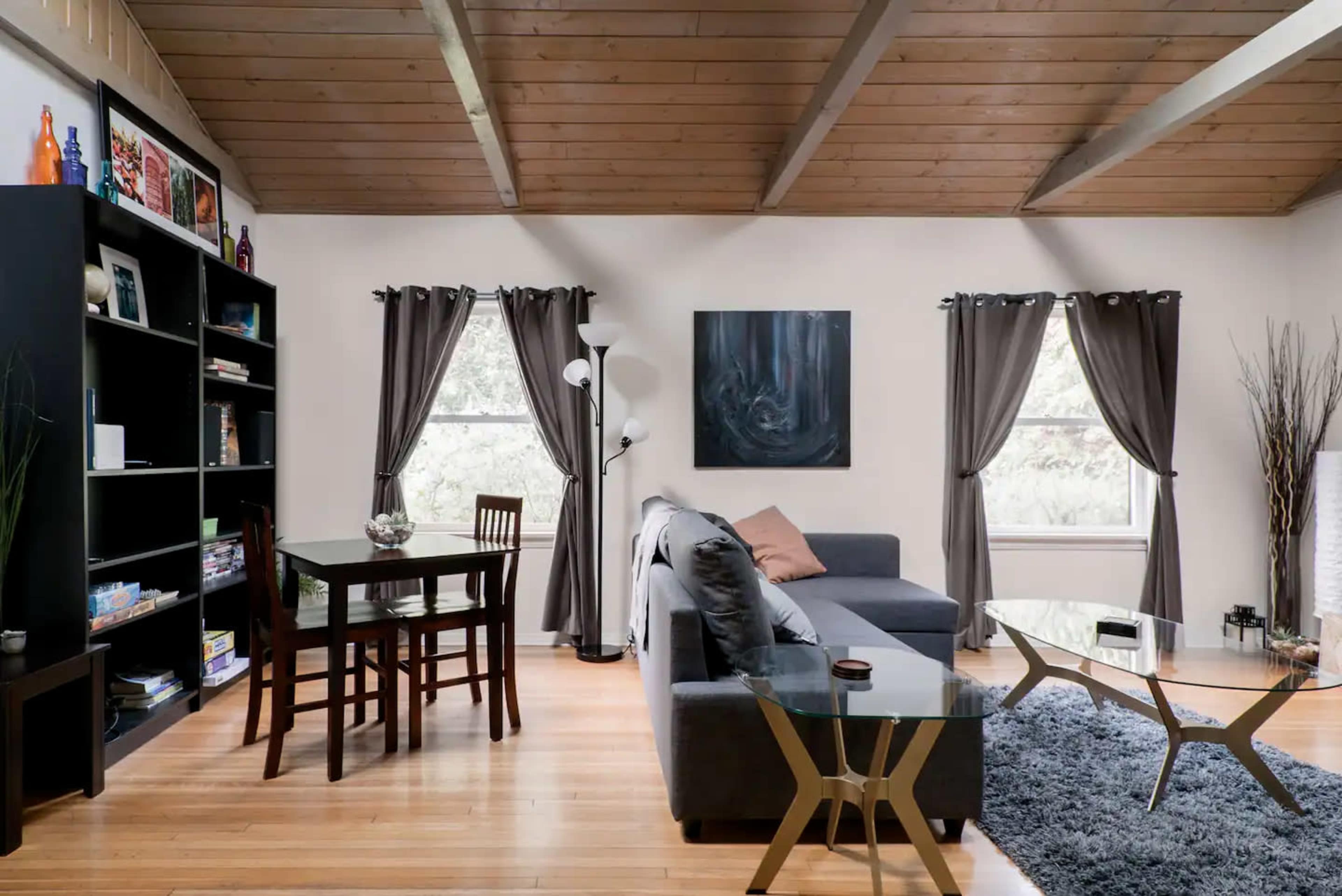 The image shows a cozy living room with a grey sofa, a wooden dining table with chairs, and a black bookshelf against the wall, featuring large windows dressed with dark curtains.