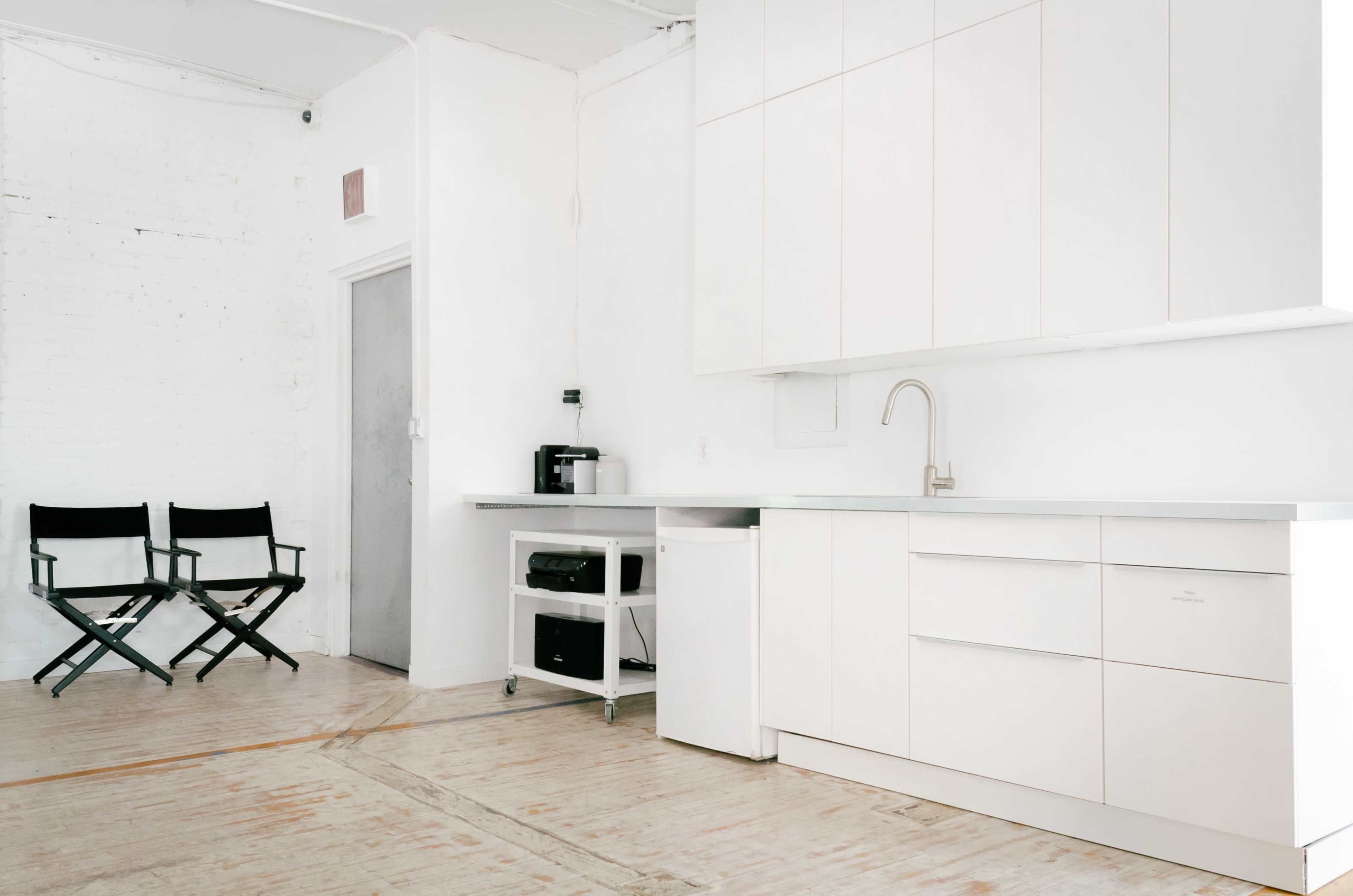 A minimalist white kitchen with a sink, countertop, and appliances, alongside two director's chairs against a white wall.
