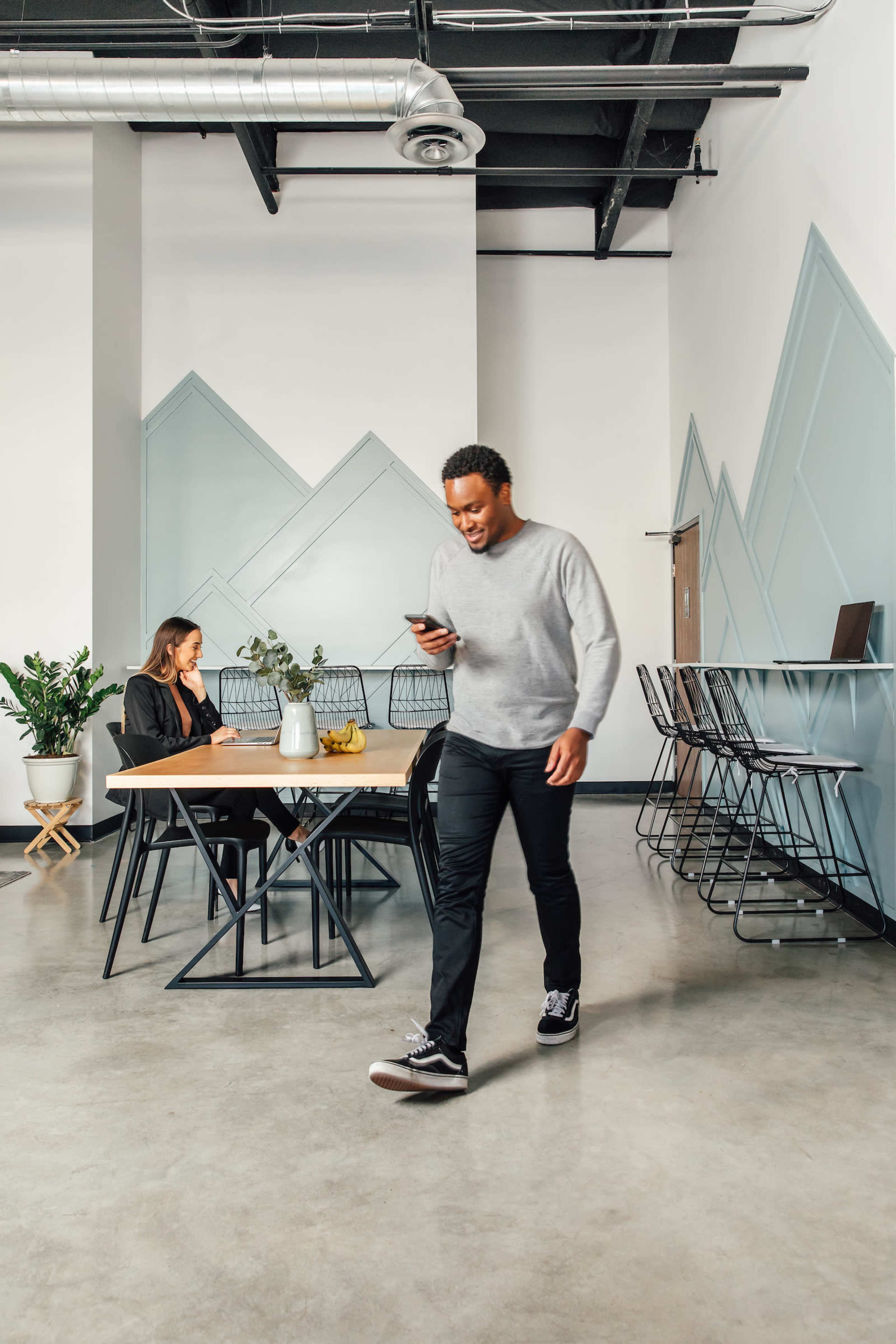 A man walks through a modern office space while a woman sits at a table, both engaged in their own activities.
