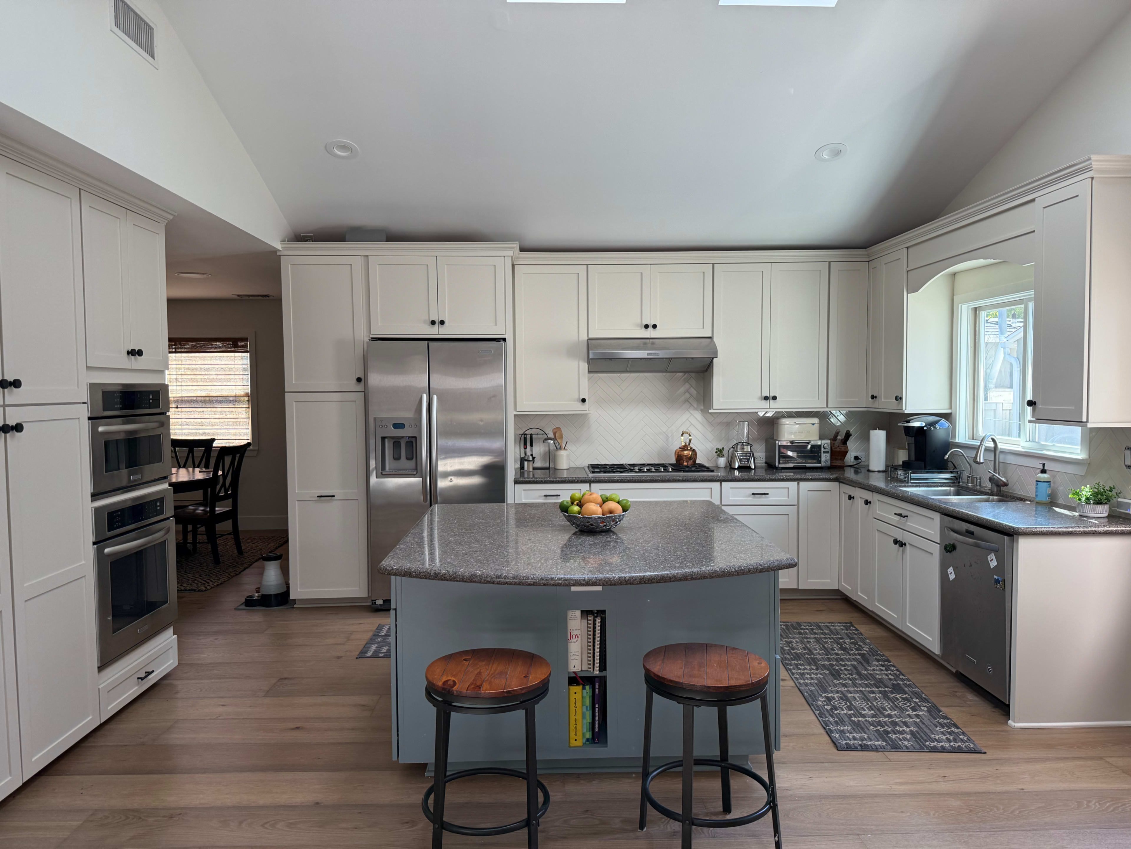 A modern kitchen features a central island with two stools, stainless steel appliances, and light-colored cabinetry.